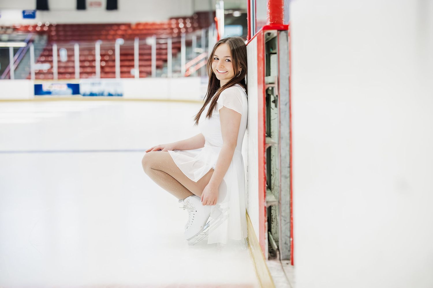 Young woman in white dress and skates posing gracefully at an indoor ice skating rink boards. Katie Chamberlain Photography