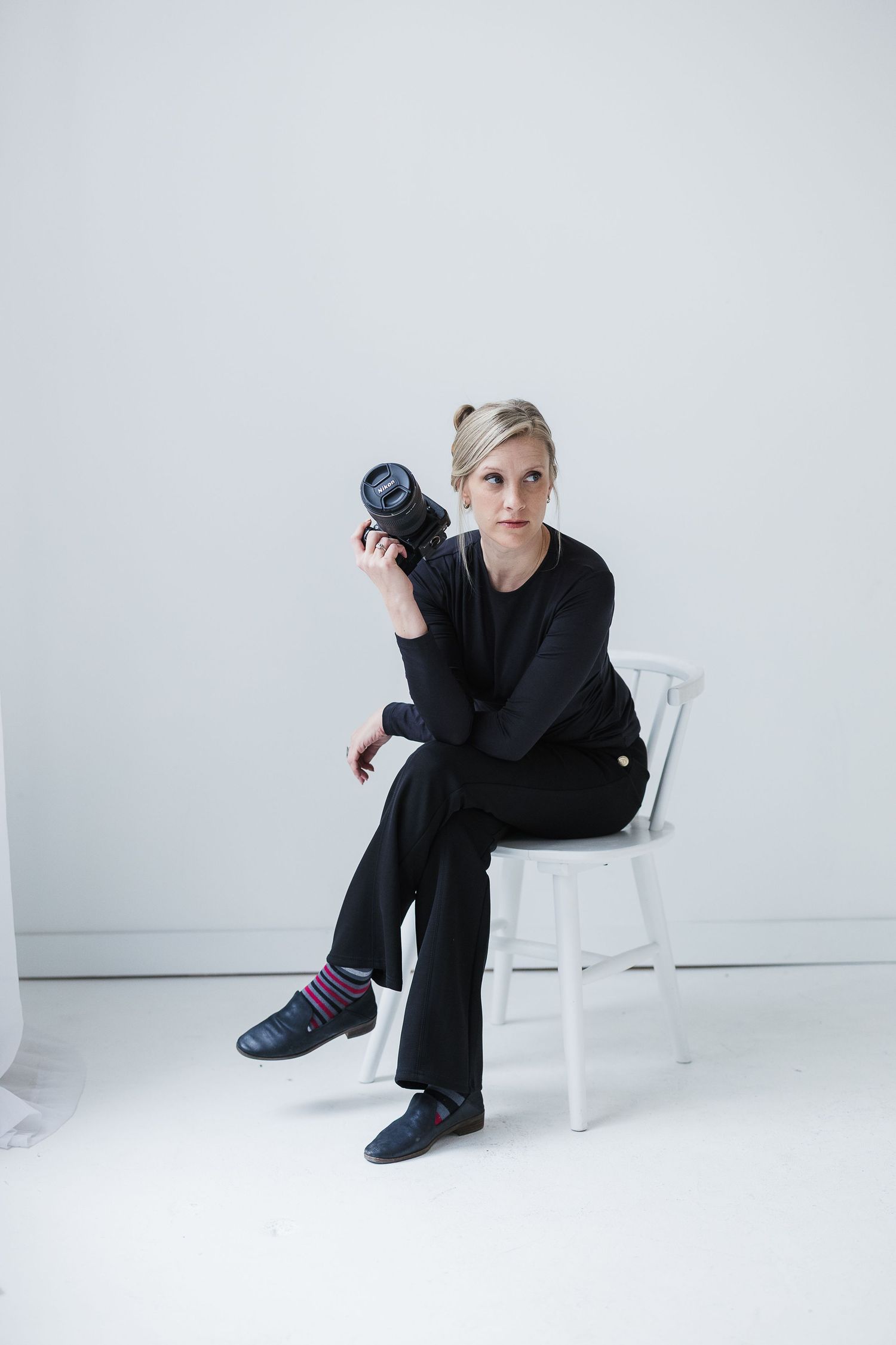 Blonde woman in black top holding a DSLR camera up to her shoulder  against a white background. Katie Chamberlain Photography