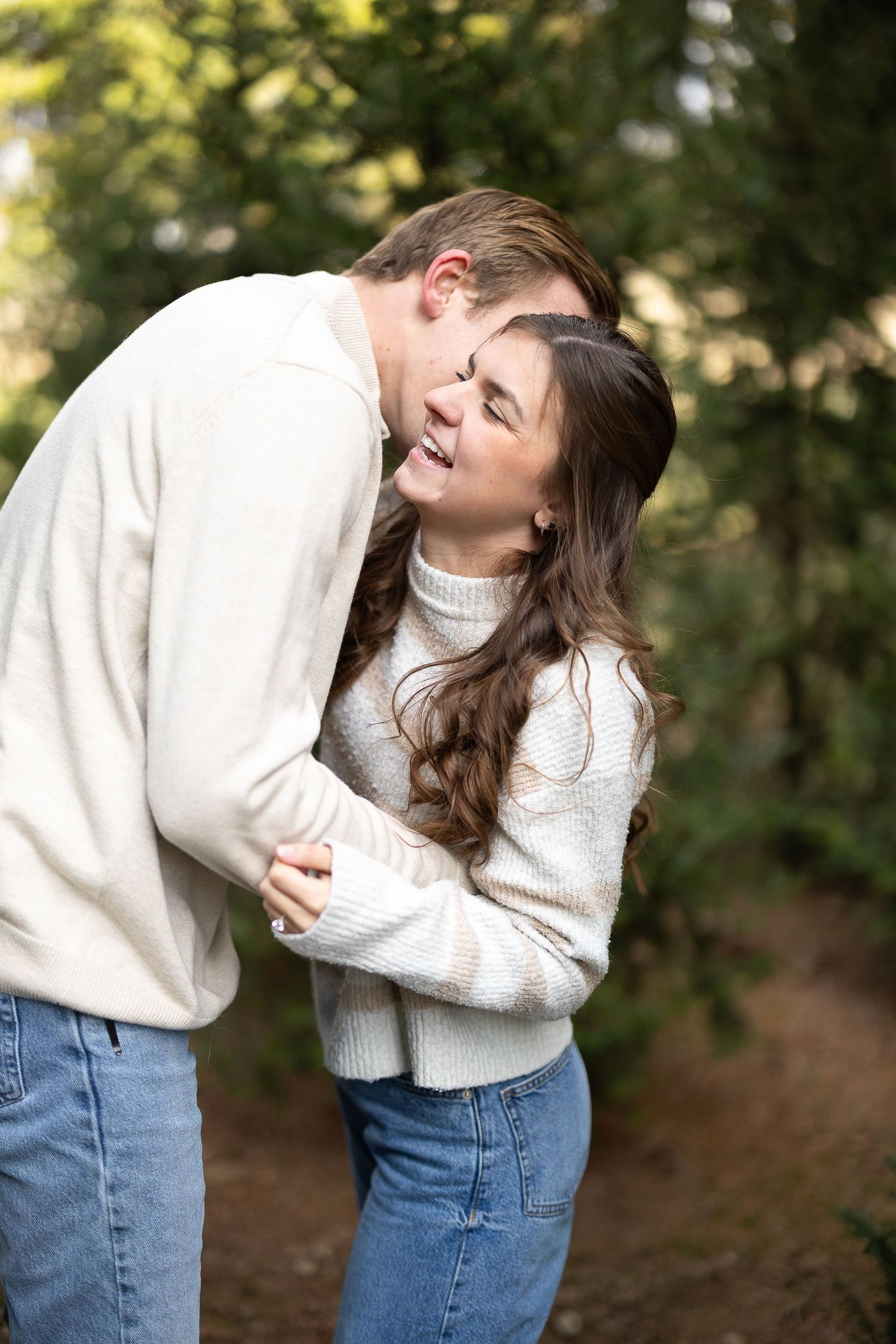 Couple in matching white sweaters and jeans embracing outdoors, woman laughing joyfully as man nuzzles her cheek. by Katie Chamberlain