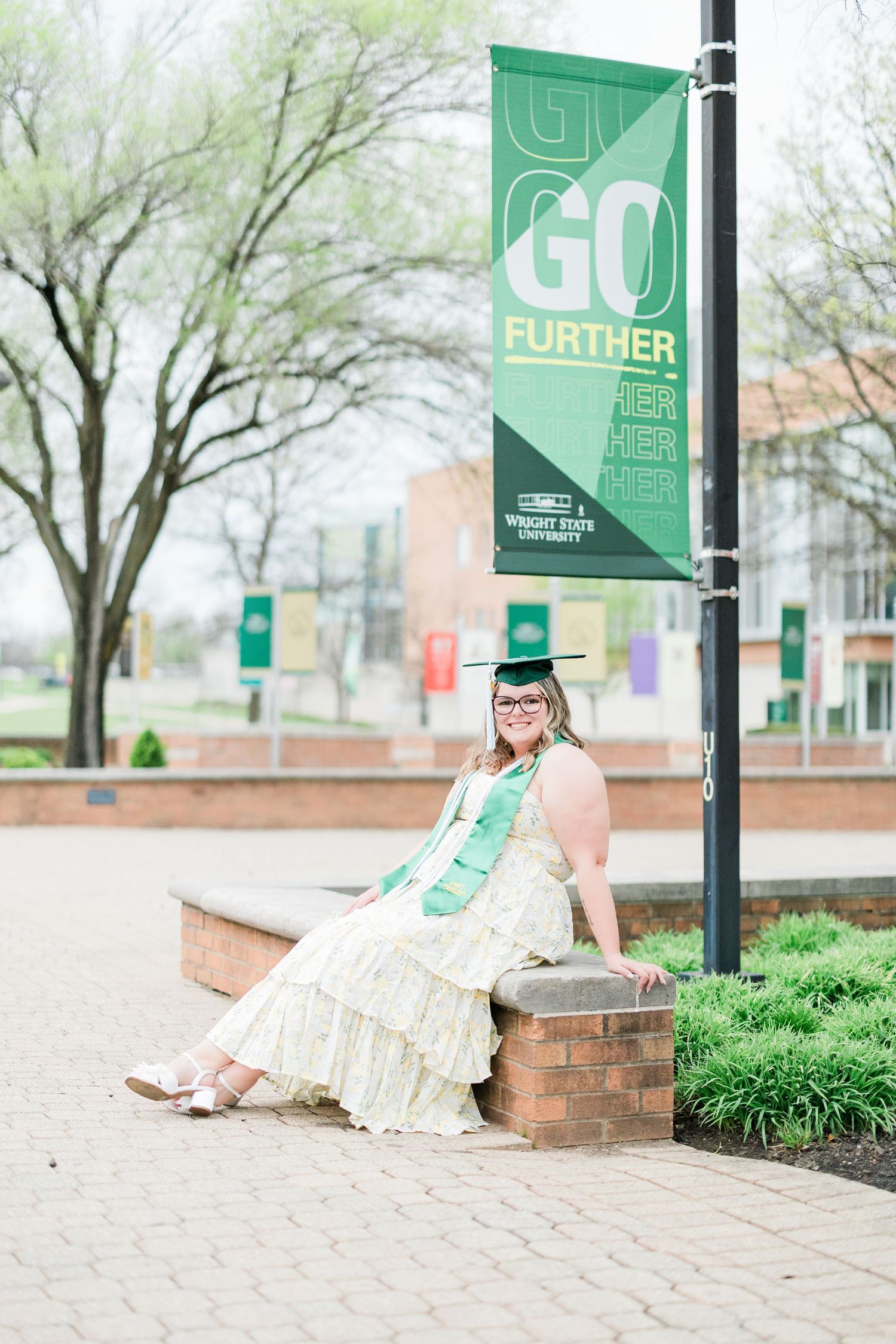 Graduate in green cap and sash sits near Wright State University Go Further banner on campus. Photographed by Katie Chamberlain Photography