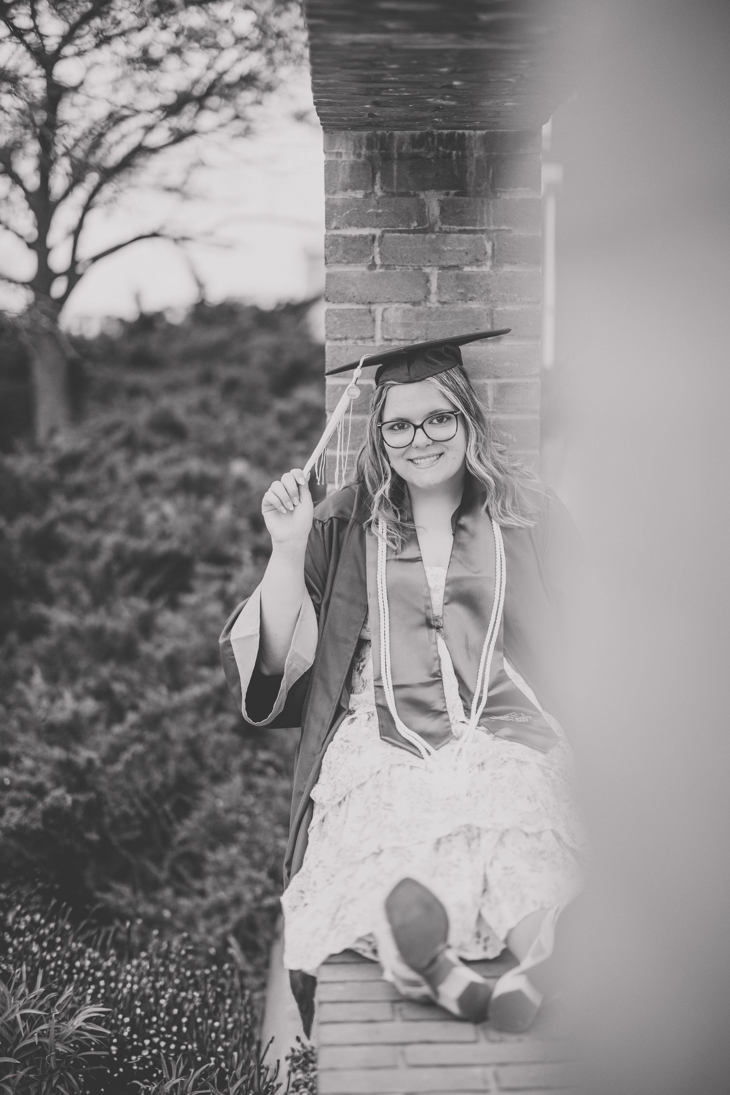Black and white photo of a graduate in cap and gown leaning against a brick column smiling. Photographed by Katie Chamberlain Photography