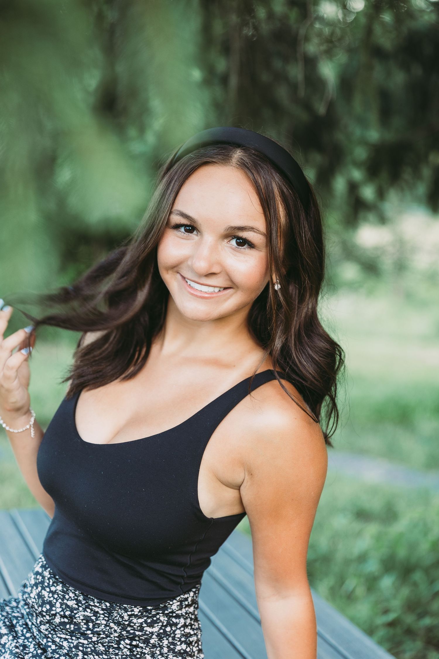 Young woman with dark hair smiling outdoors in a black tank top and patterned skirt near green trees. Katie Chamberlain Photography