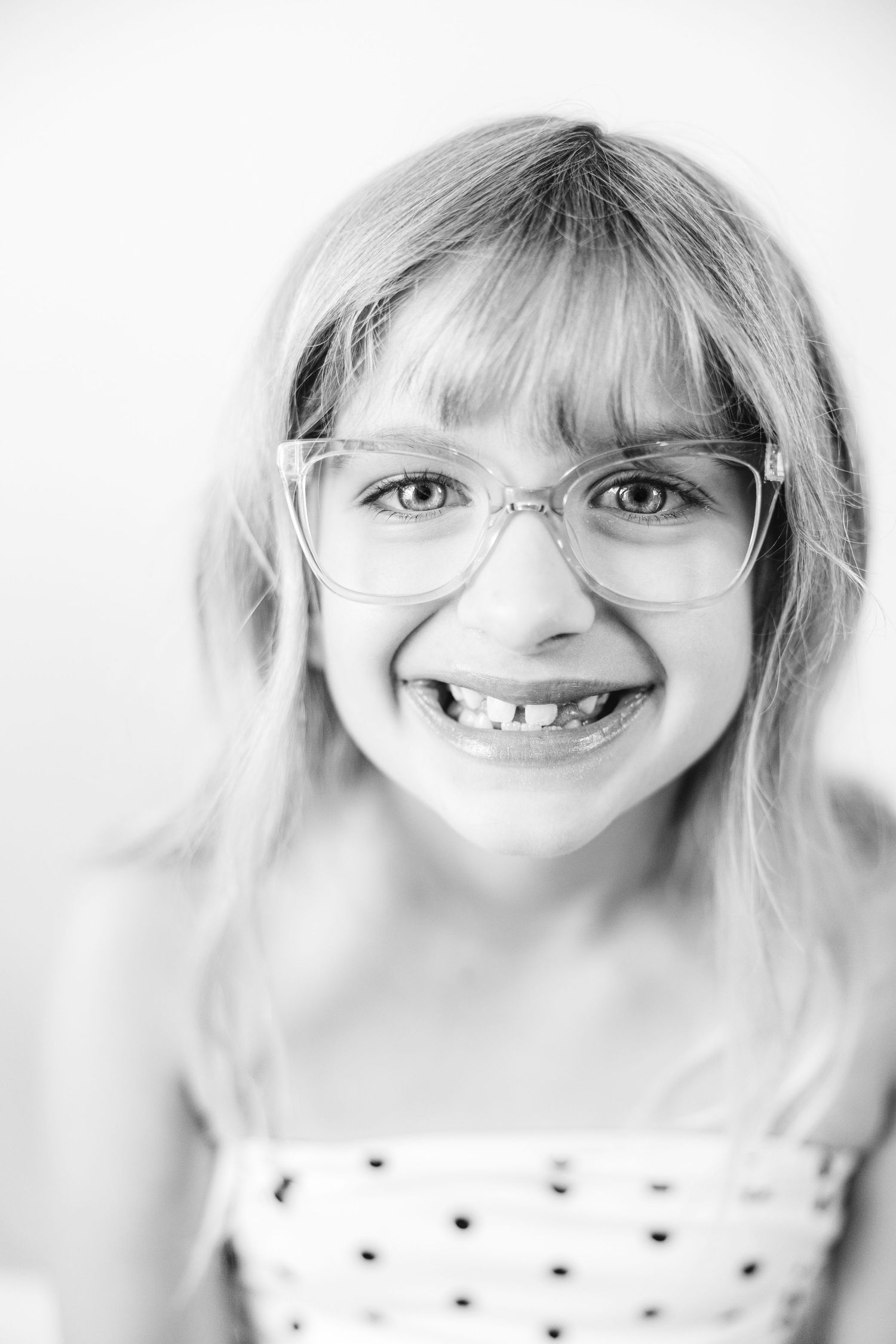 Black and white portrait of a smiling young girl with glasses and long blonde hair showing her missing teeth.