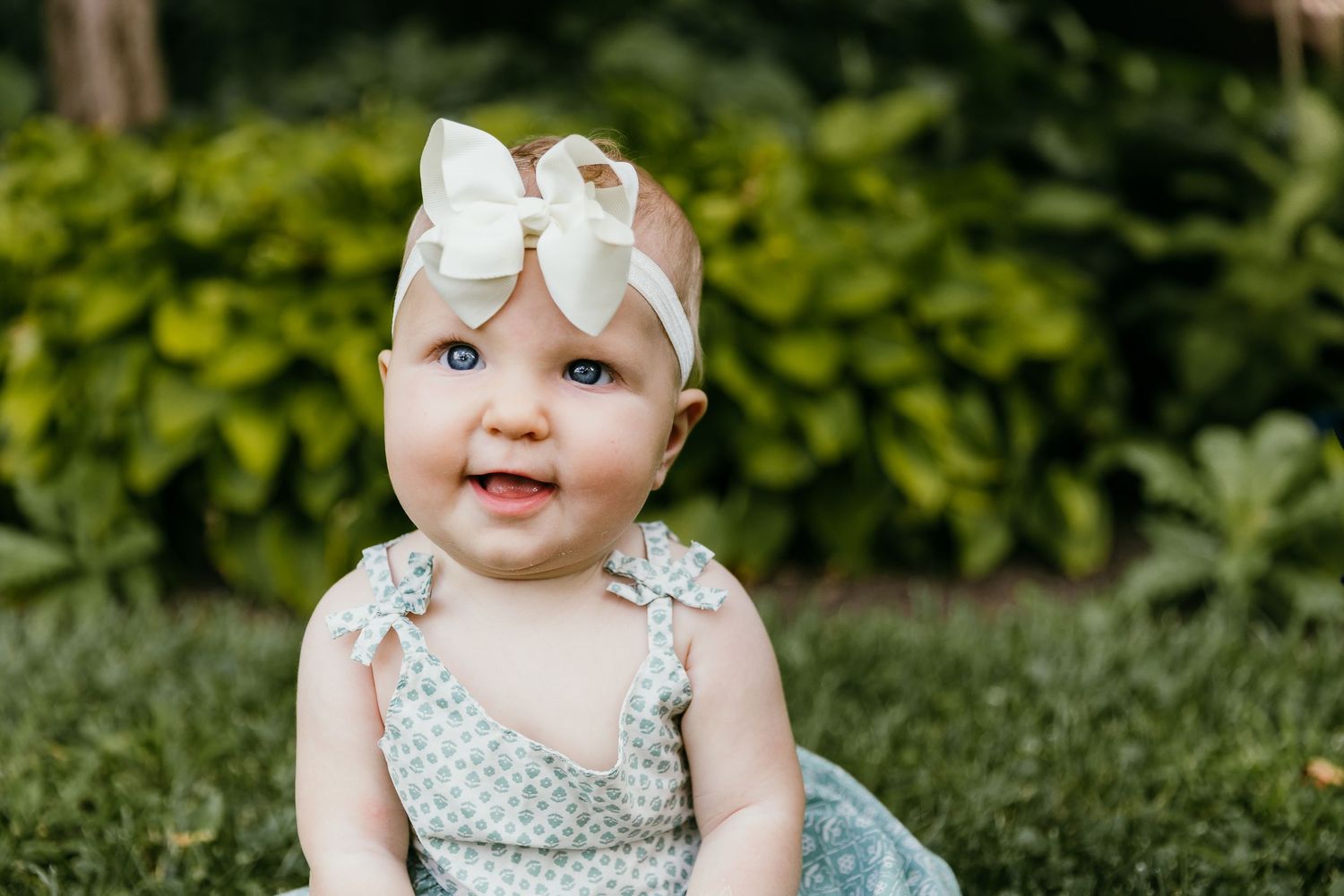 Adorable baby girl with blue eyes wearing a floral romper and white bow headband sitting on green grass. Photographed by Katie Chamberlain