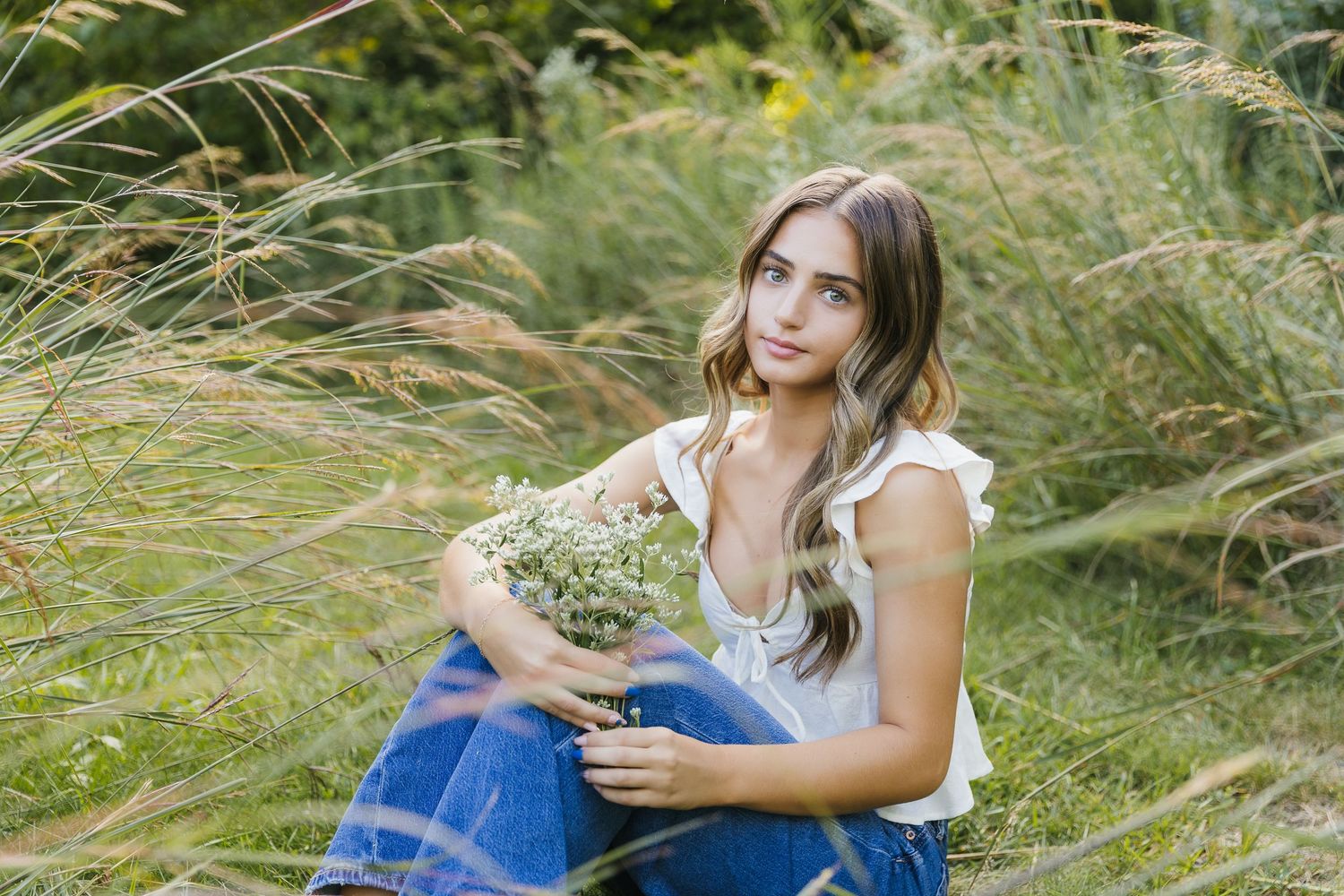 Young woman sitting in tall grass holding wildflowers, wearing white top and jeans in a serene outdoor setting. Katie Chamberlain Photograph