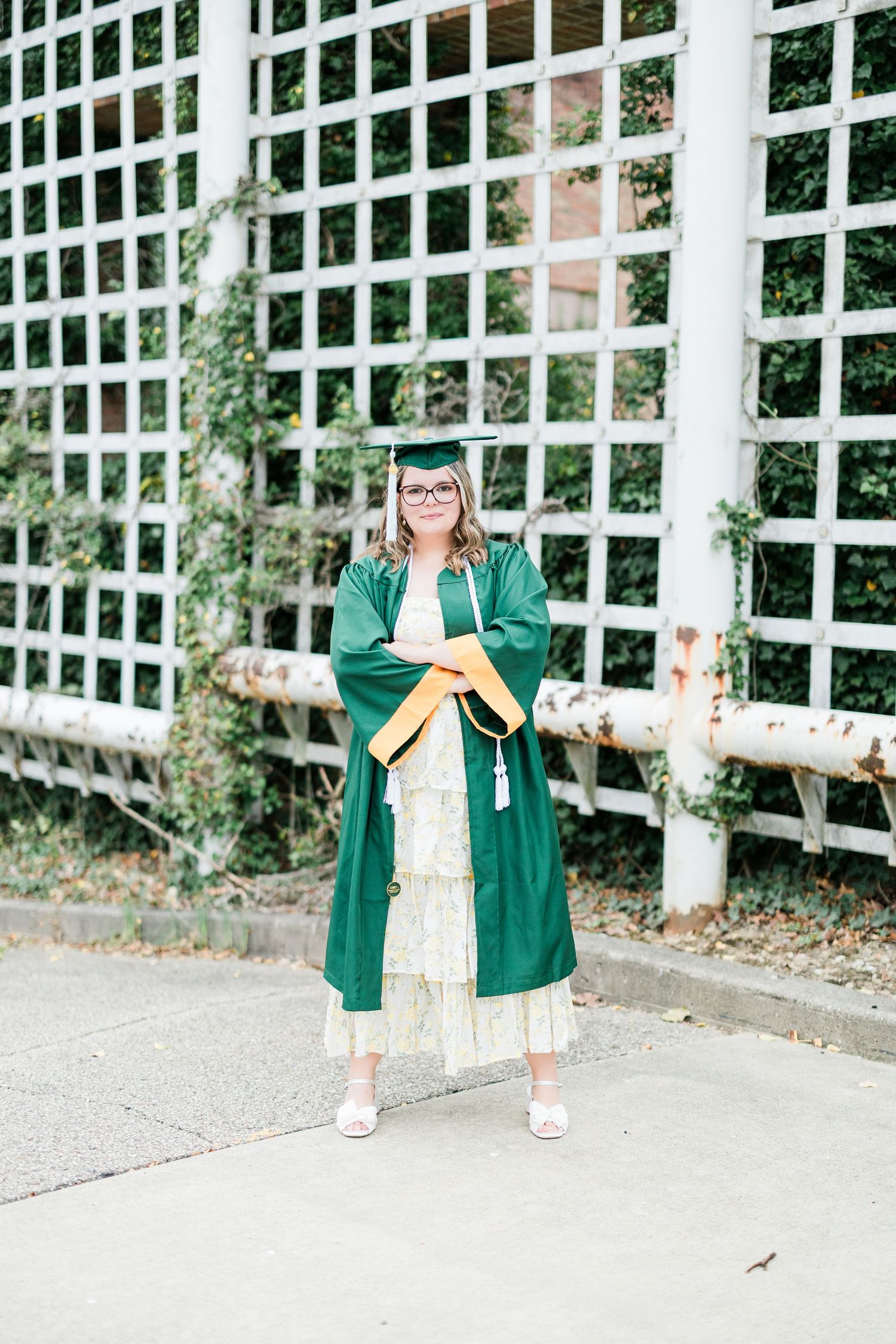 Young woman in green graduation gown and cap stands proudly outdoors near white lattice fence. Photographed by Katie Chamberlain Photography