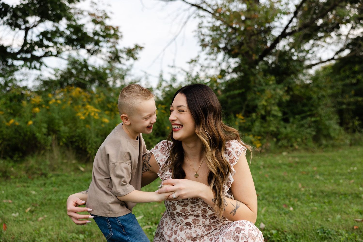 A joyful mother in a floral dress laughs with her young blond son outdoors in a lush green park setting.