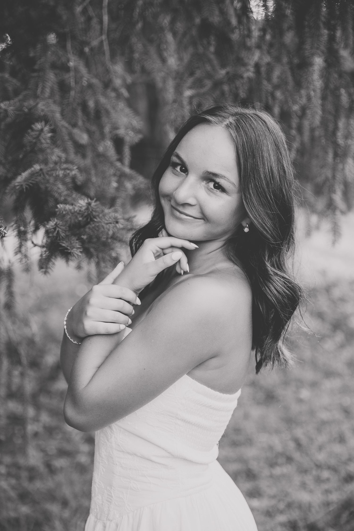 Black and white portrait of a young woman in a strapless white dress smiling outdoors near pine trees. Katie Chamberlain Photography