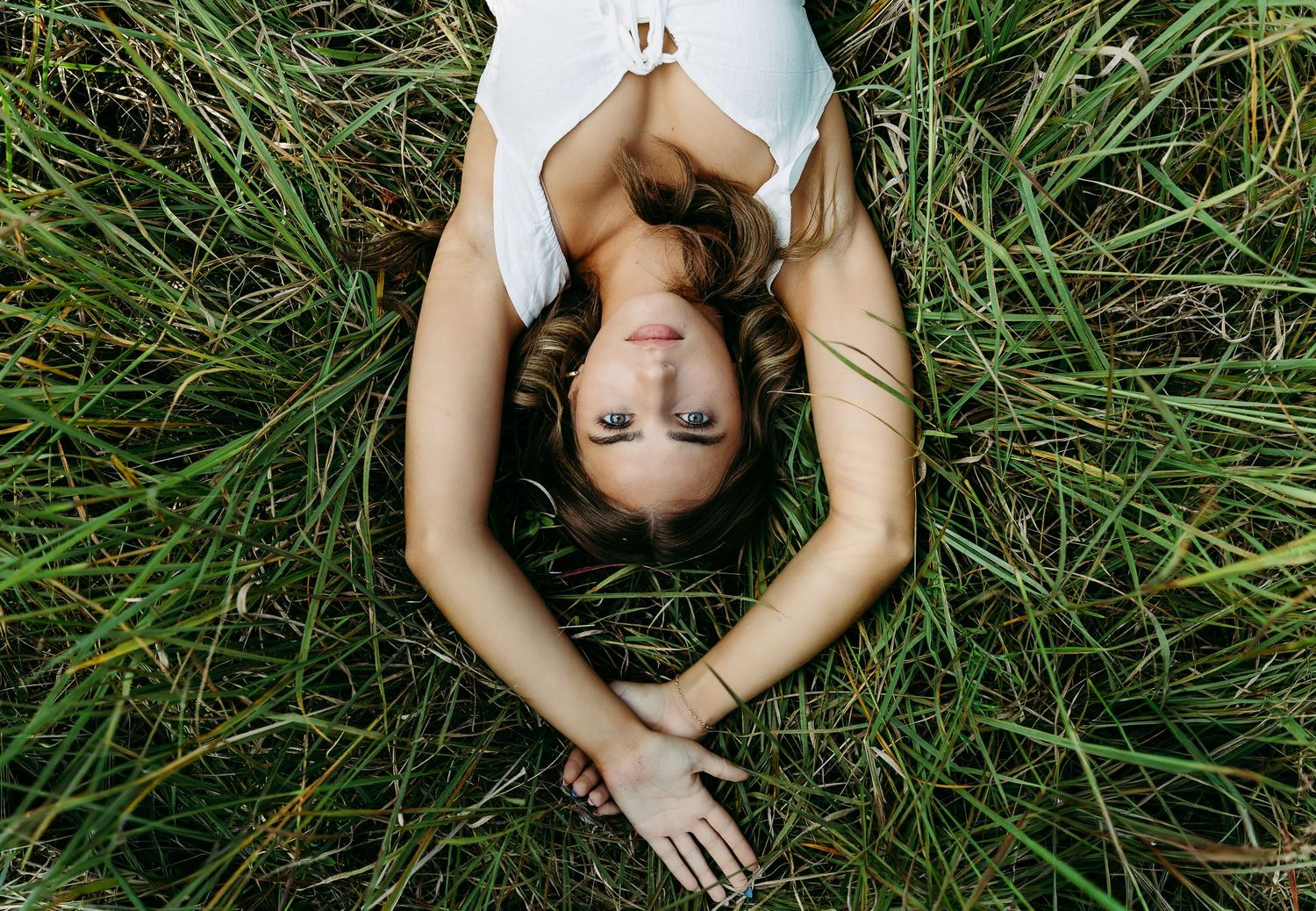 Young woman lying in green grass, looking up at the sky, wearing a white top on a sunny day. Photographed by Katie Chamberlain Photography