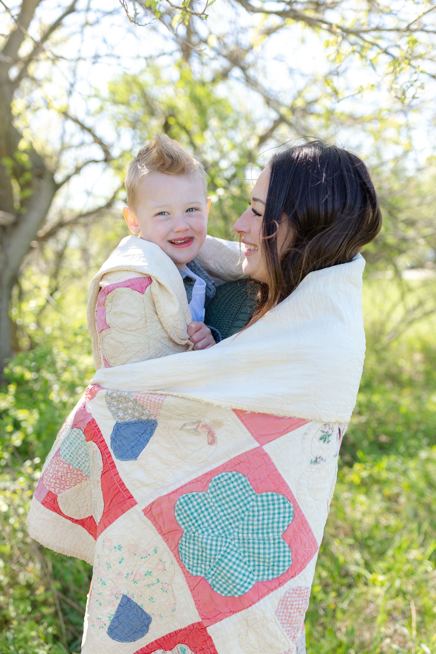 A mother wraps her smiling baby in a colorful patchwork quilt outdoors among green trees on a sunny day.