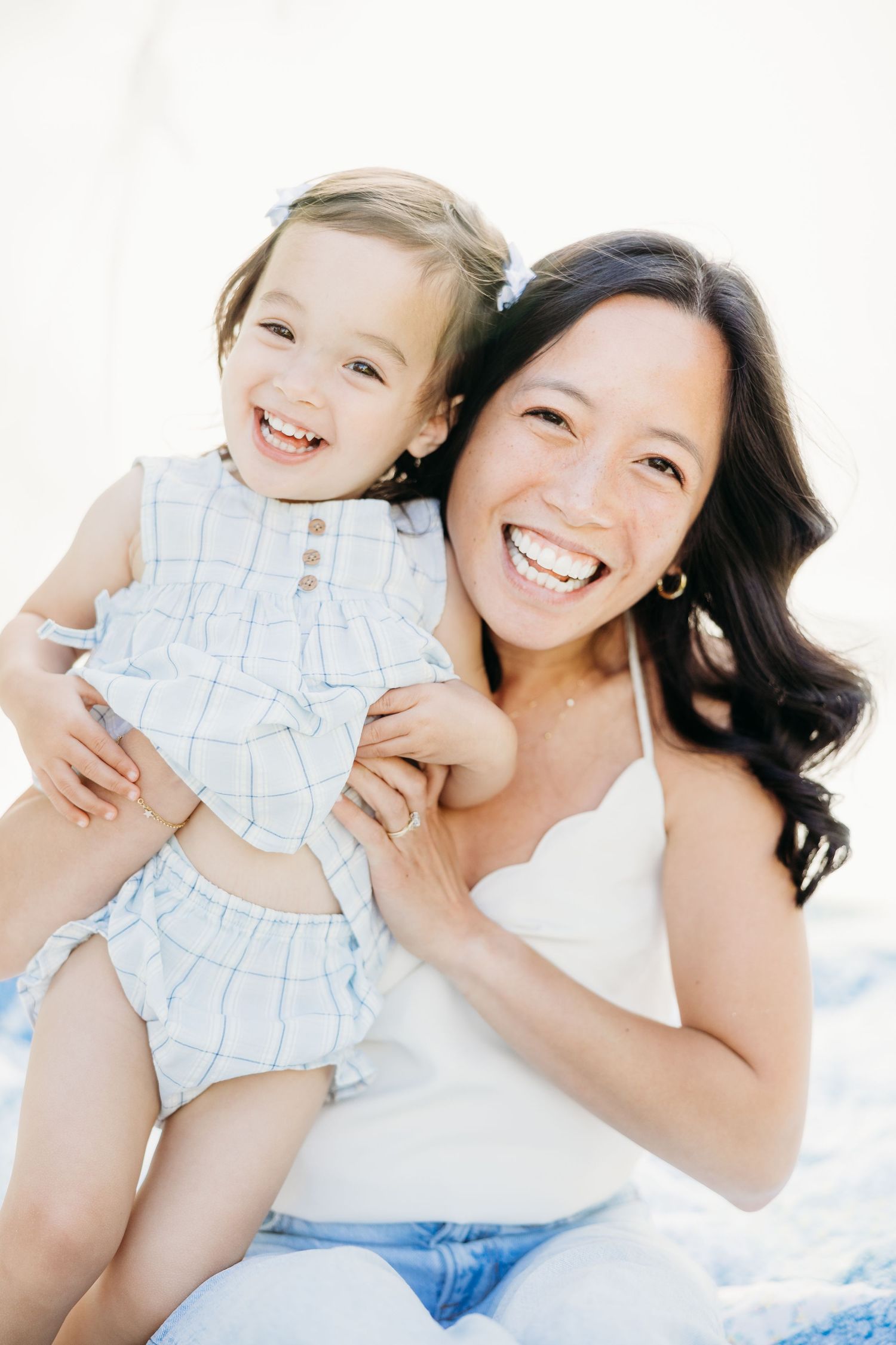 A smiling woman in white holds a laughing toddler girl in a light blue outfit against a bright background.