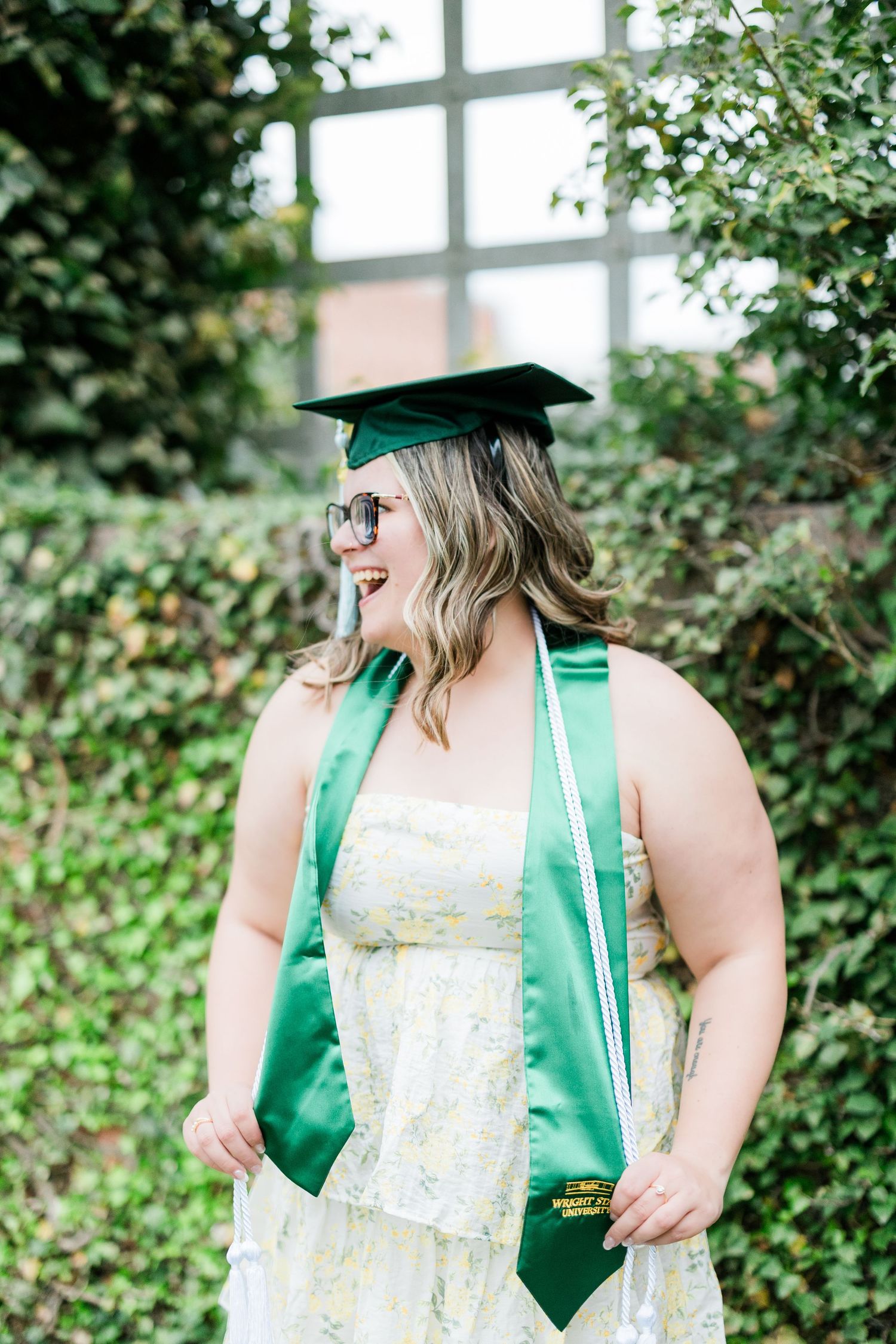 Smiling graduate wearing green cap and sash with sunglasses poses in front of ivy-covered wall. Photographed by Katie Chamberlain Photograph
