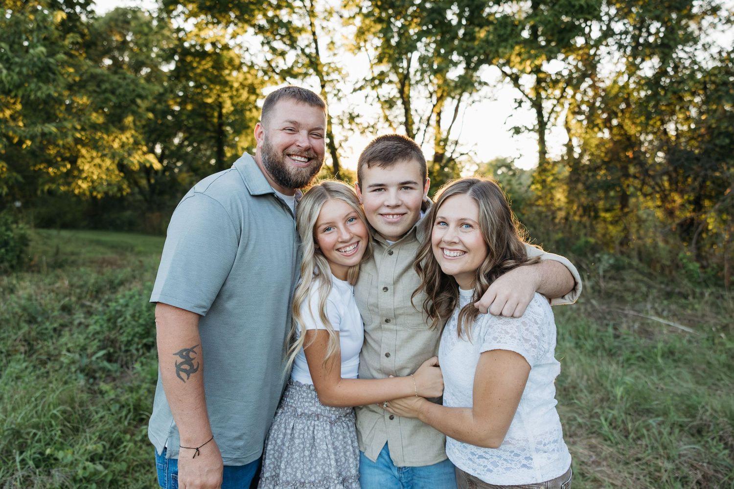 Happy family of four smiling together outdoors in a lush green field during golden hour.