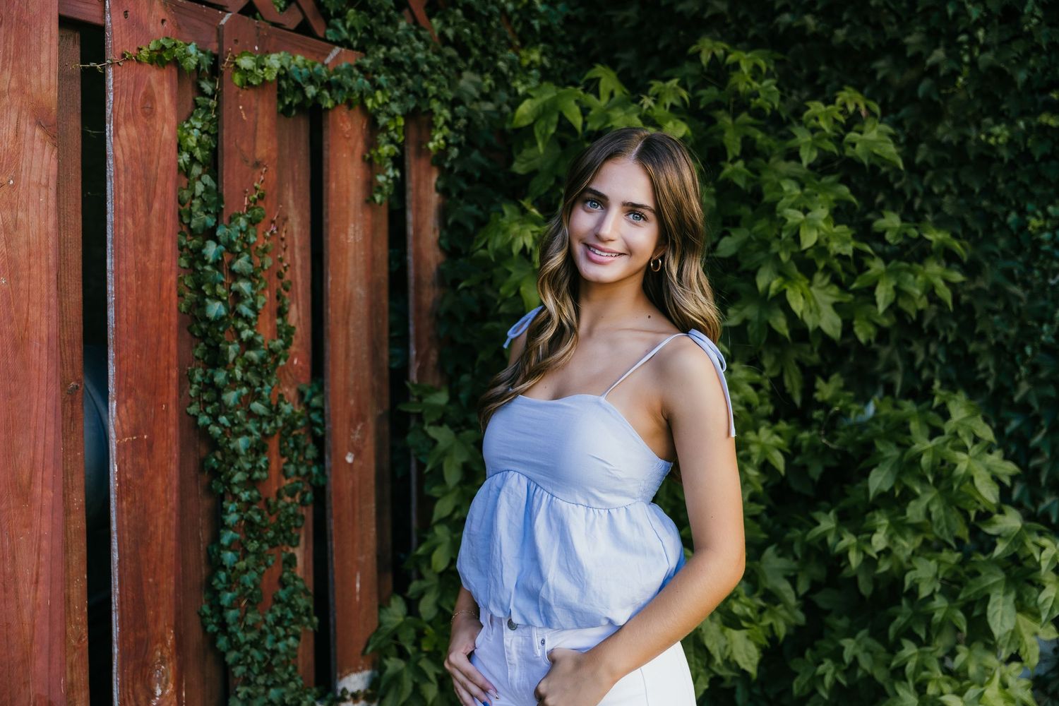 Smiling pregnant woman in blue top and white skirt standing by ivy-covered wooden fence outdoors. Katie Chamberlain Photography