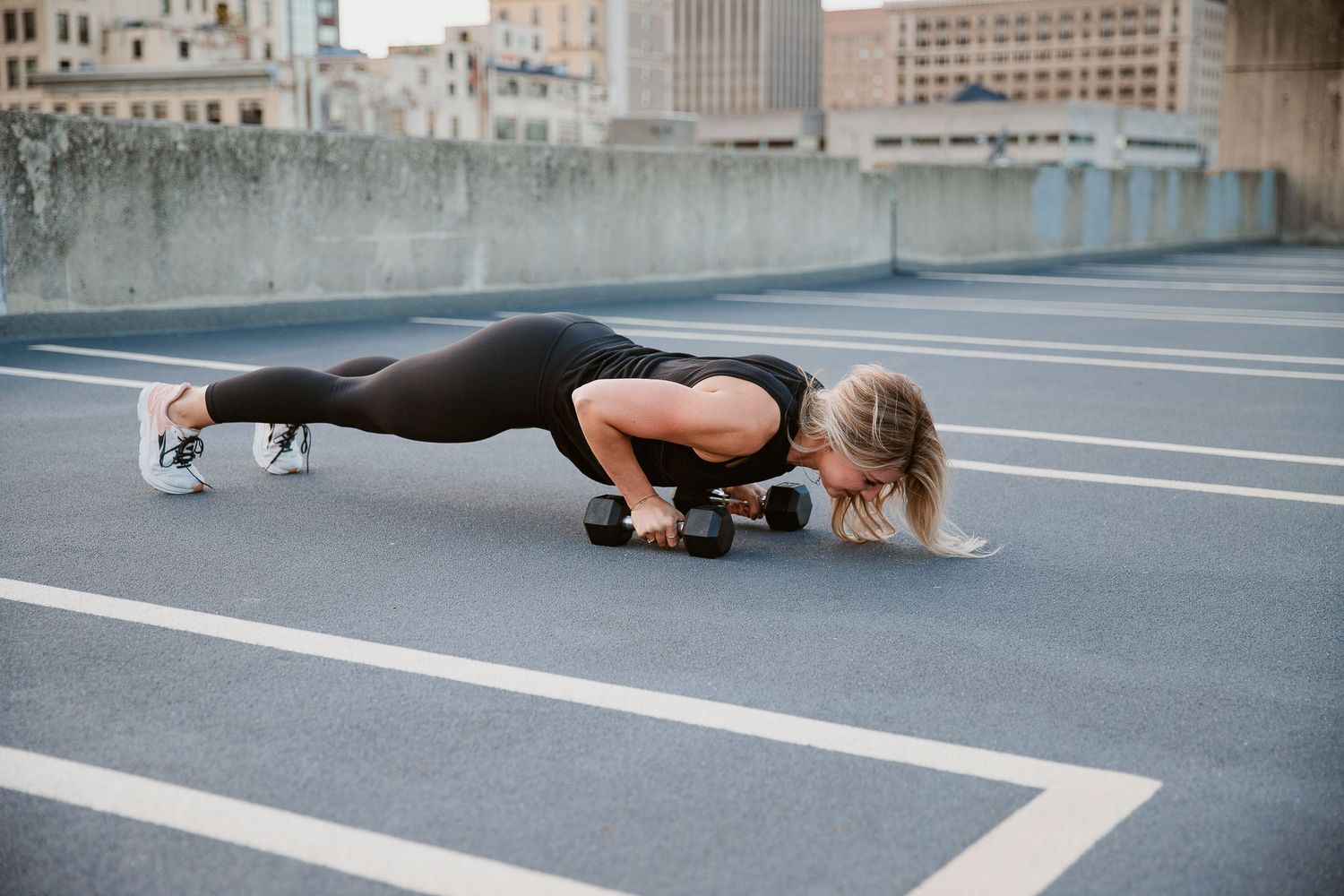 Woman in black athletic wear doing push-ups on dumbbells on a rooftop parking lot in an urban setting. Photographed by Katie Chamberlain