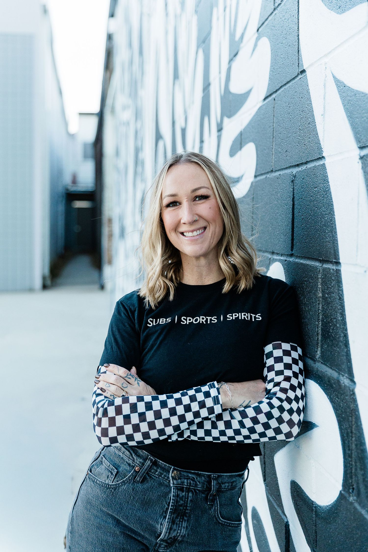 Smiling blonde woman in black Subs Sports Spirits t-shirt with checkered sleeves, arms crossed, posing in urban alley. Katie Chamberlain