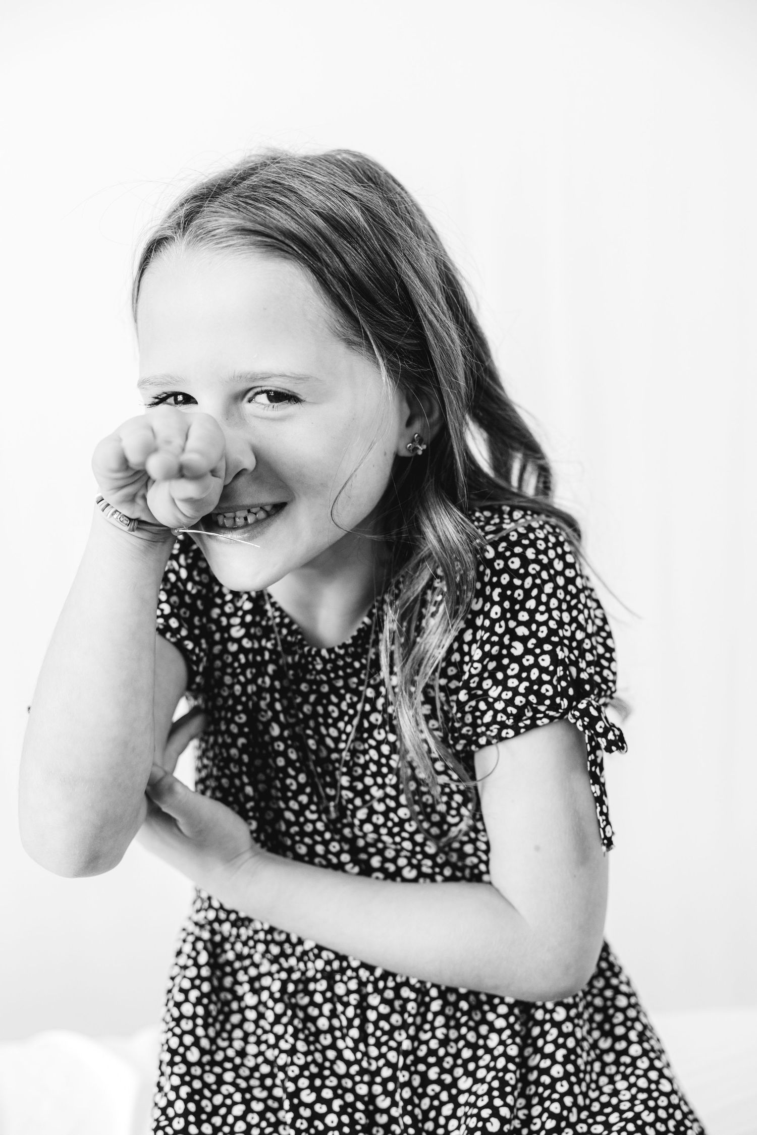 Black and white portrait of a smiling young girl in a floral dress, playfully covering her mouth with her hand.