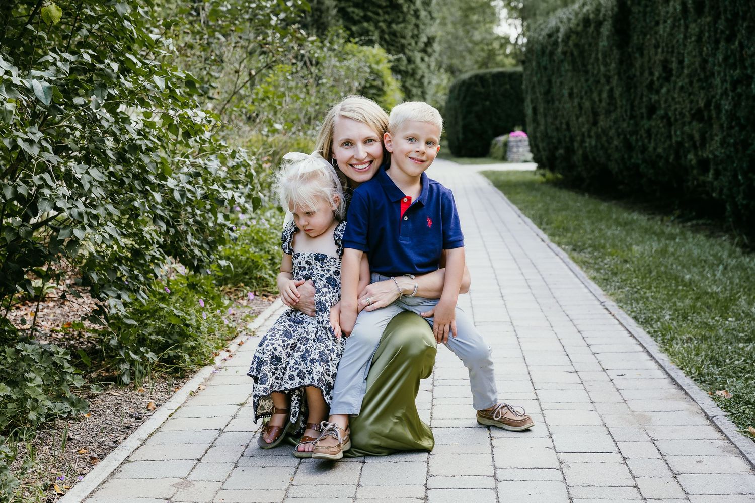 A smiling woman crouches on a brick path with two young children, surrounded by green hedges outdoors. Photographed by Katie Chamberlain