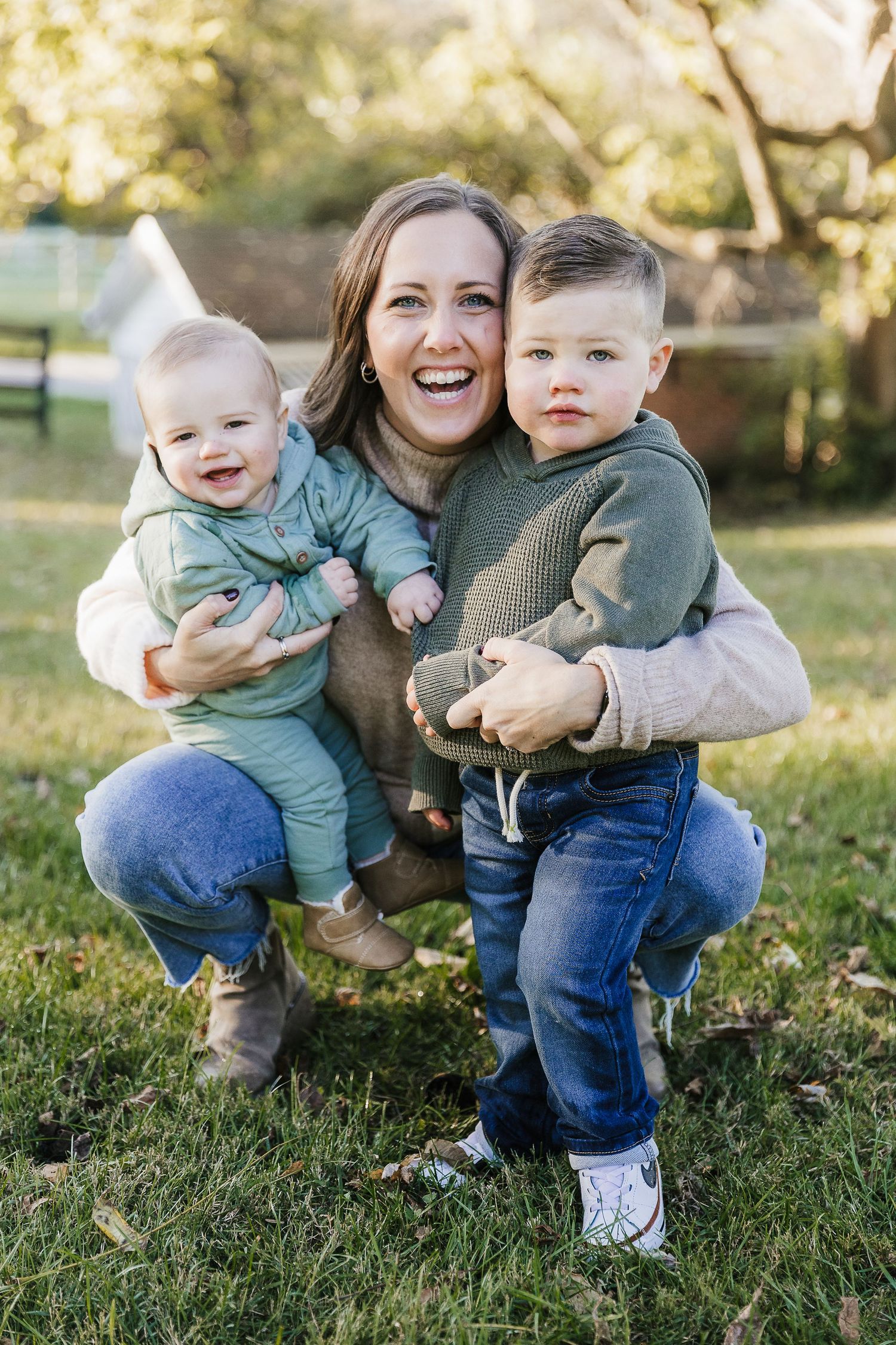 Smiling woman crouching outdoors holding two young toddler boys in green hoodies and jeans on a sunny day. Photographed by Katie Chamberlain