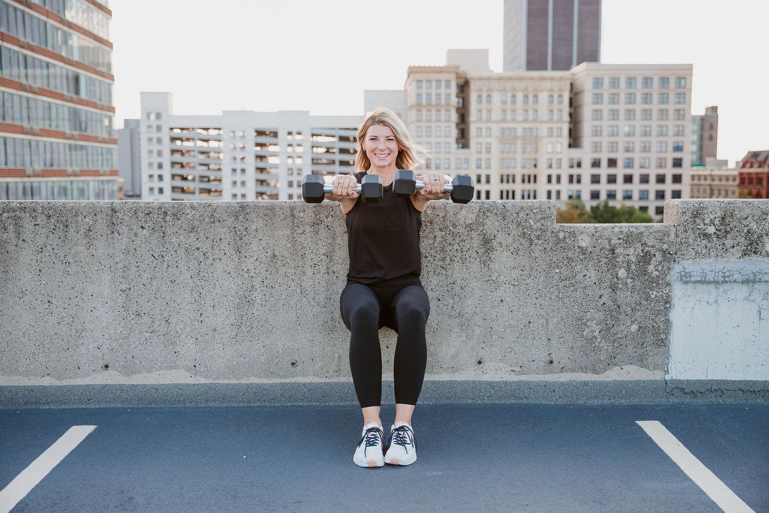 Smiling blonde woman in black athletic wear leaning against a concrete wall urban buildings in the background. Photo by Katie Chamberlain