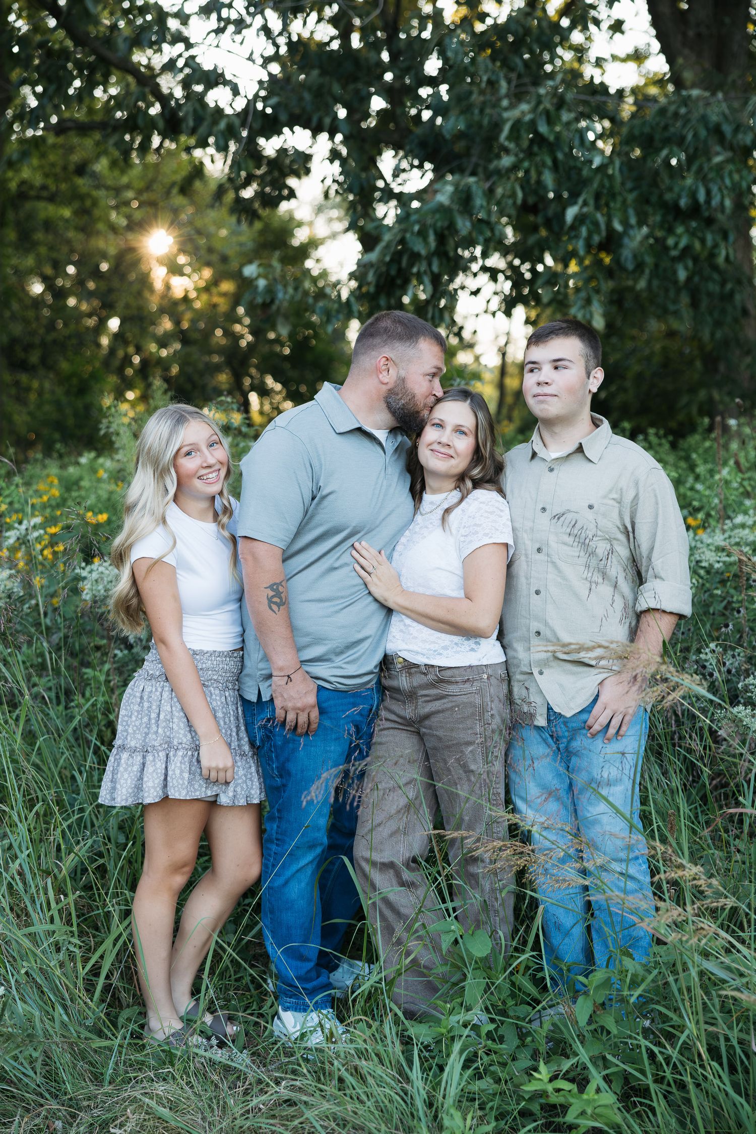 Family of four posing outdoors in a lush green field at golden hour, wearing casual earth-toned outfits.
