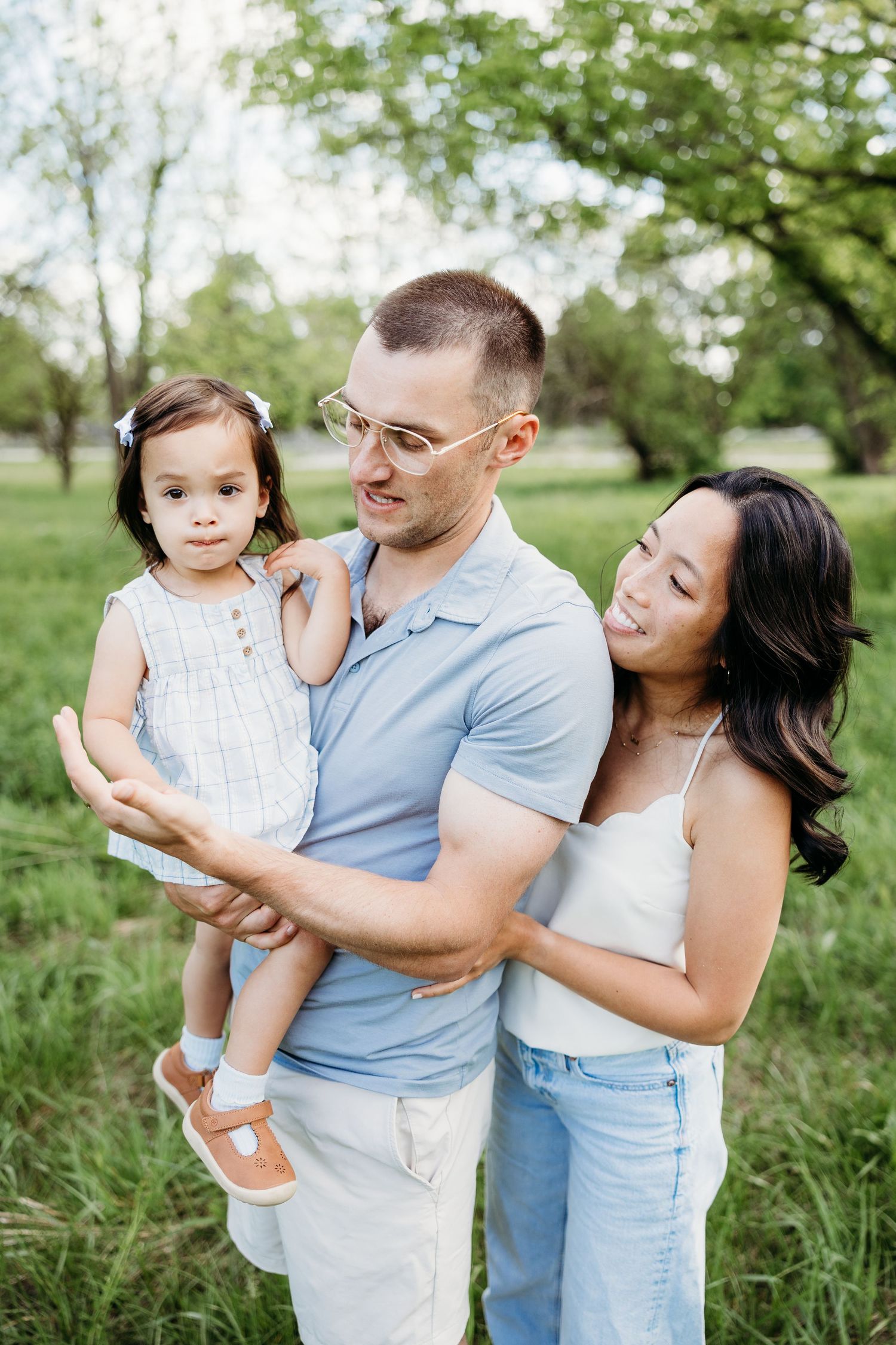 A young family with a toddler girl poses outdoors in a green park, parents smiling lovingly at their child.