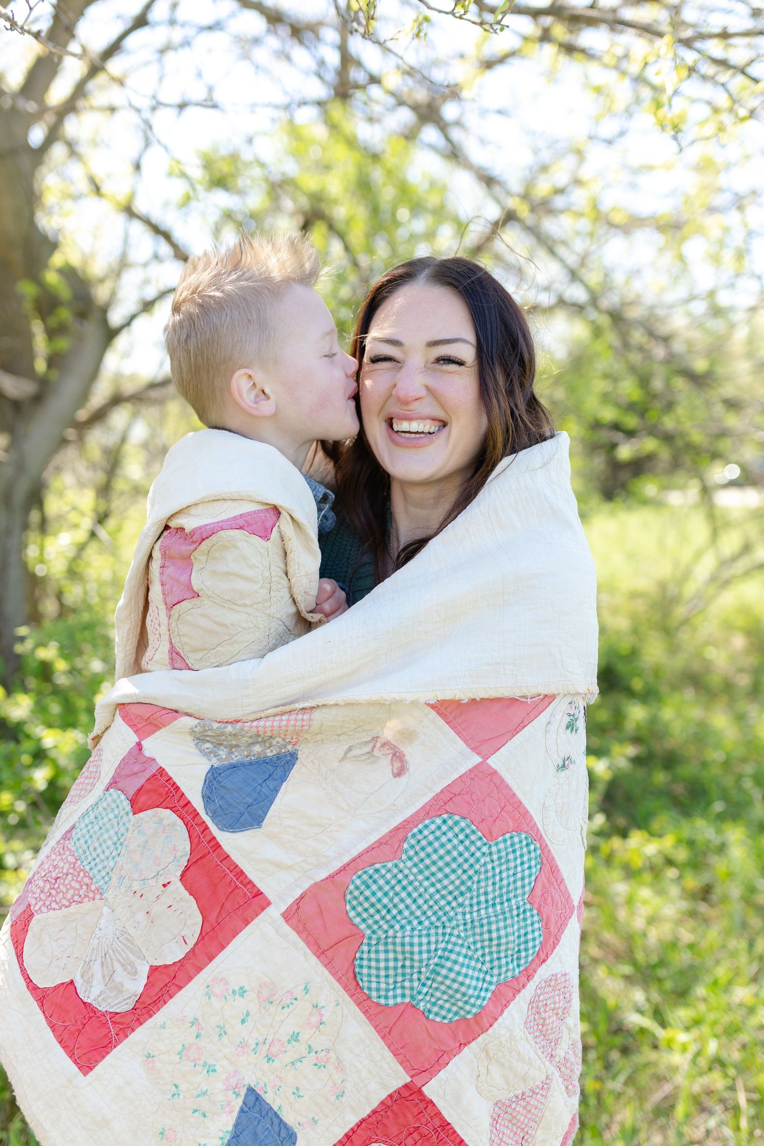 A laughing mother and young child wrapped in a colorful patchwork quilt outdoors, sharing a sweet kiss.