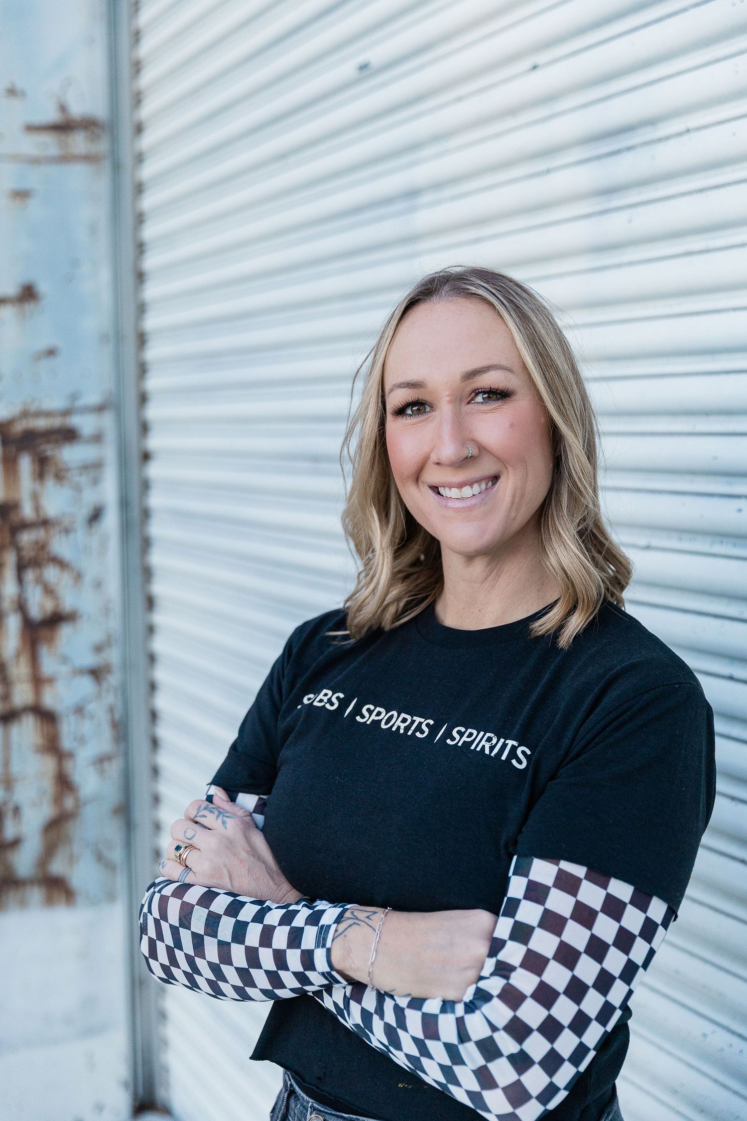Smiling blonde woman in black Subs Sports Spirits t-shirt with checkered sleeves, arms crossed, posing in urban alley. Katie Chamberlain