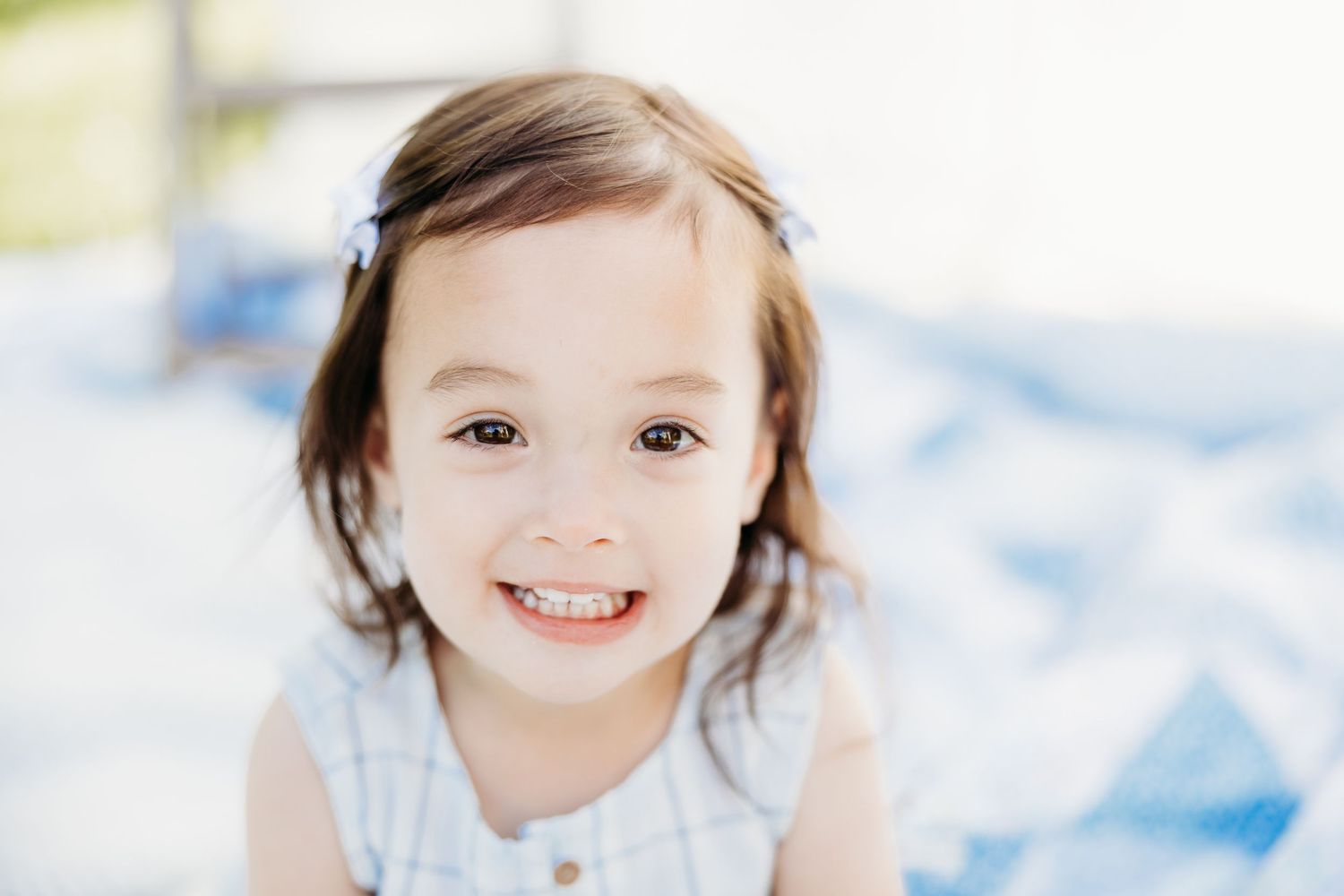 Smiling toddler girl with hair clips wearing a white dress, sitting outdoors on a blue blanket.