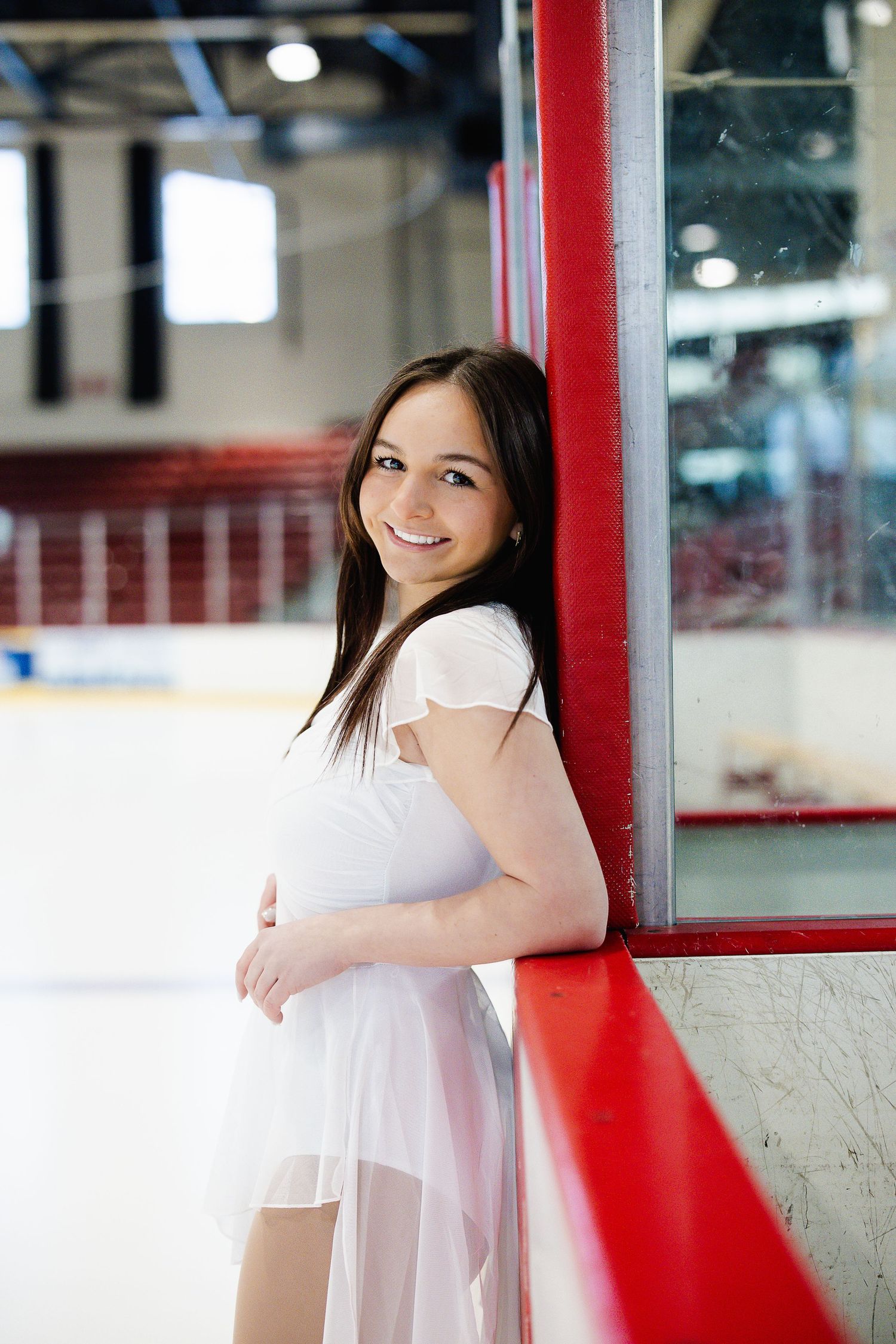 Young woman in white dress smiling at ice rink boards, figure skating arena background. Katie Chamberlain Photography