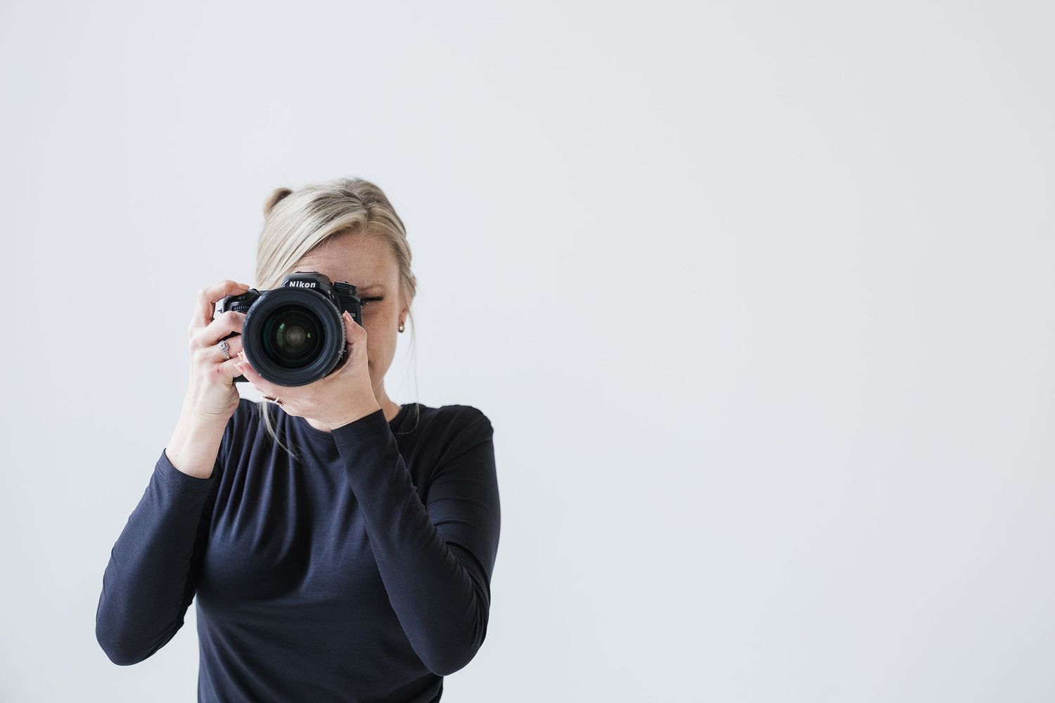 Blonde woman in black top holding a DSLR camera up to her eye against a white background. Katie Chamberlain Photography
