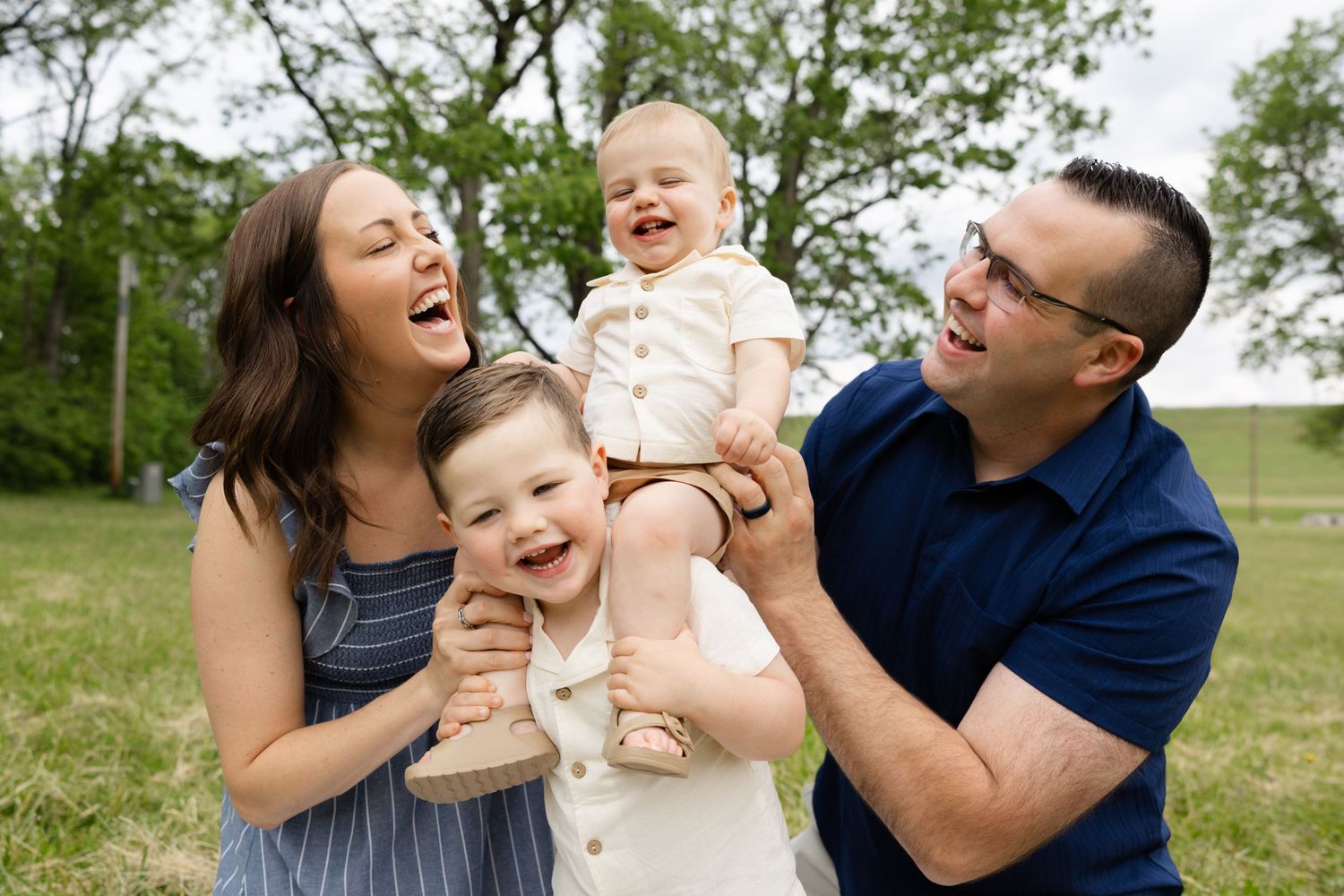 Happy family of four laughing outdoors, parents holding two young children in a green park setting.