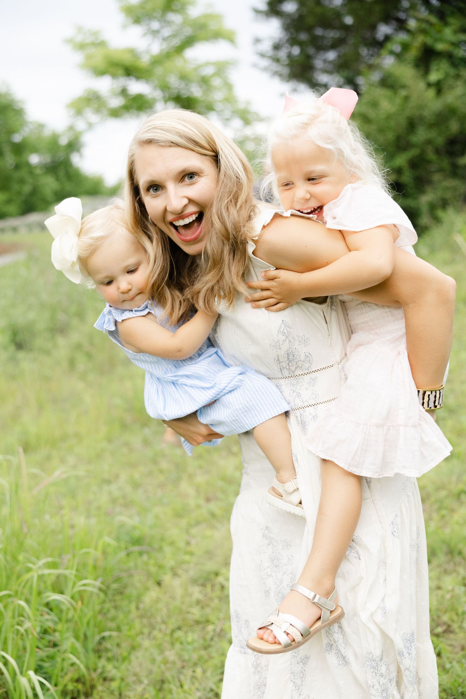 A joyful blonde mother in a white dress holds two young daughters outdoors in a green field.