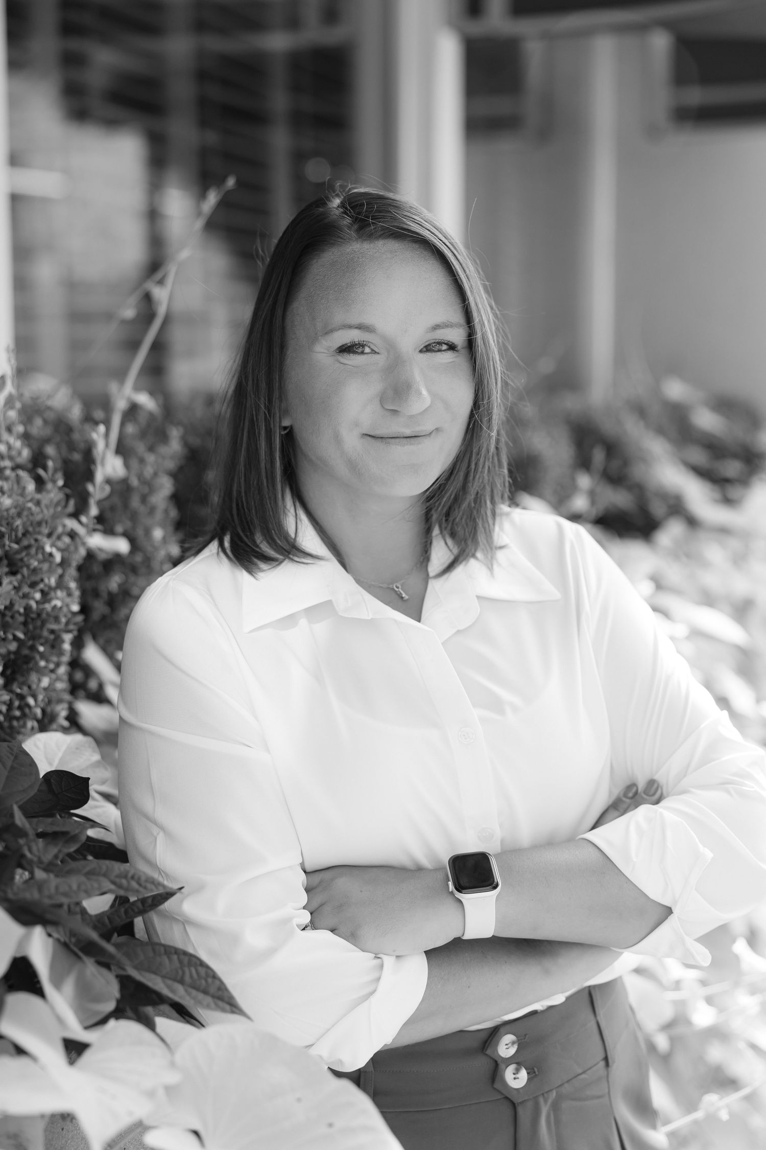 Black and white portrait of a smiling woman in white shirt, arms crossed, wearing smartwatch, outdoors with plants.
