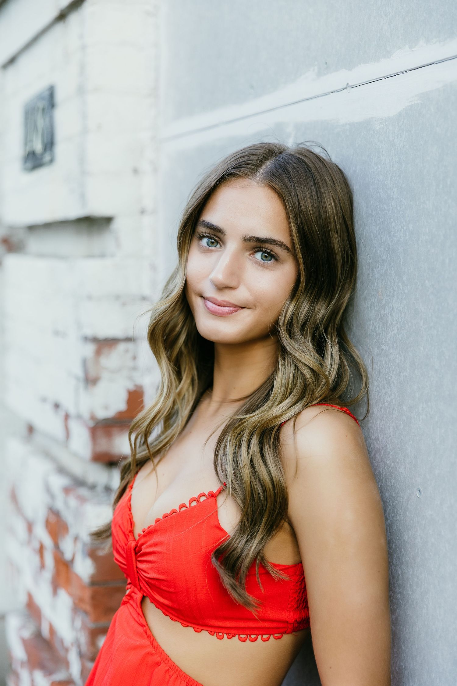 Young woman with wavy brown hair wearing a red crop top, leaning against a gray wall outdoors. Katie Chamberlain Photography