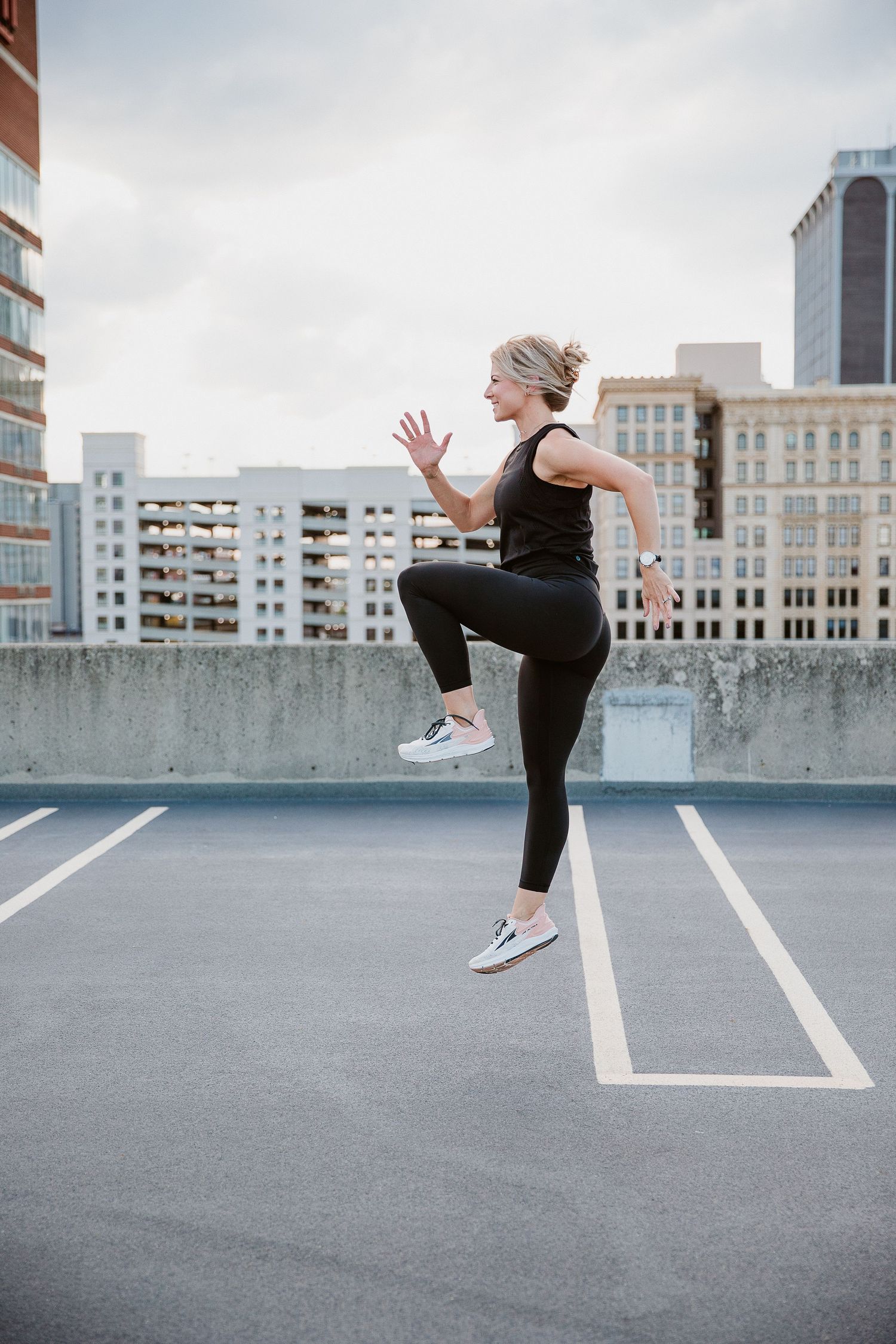 Woman in black athletic wear doing high knees exercise on rooftop parking lot with city skyline background. Photo by Katie Chamberlain