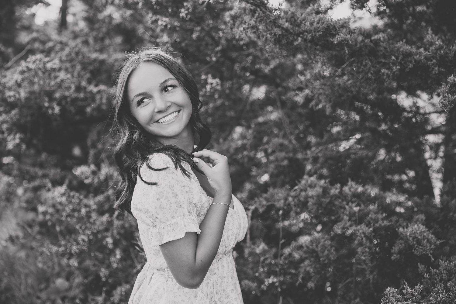 Black and white portrait of a smiling young woman in a floral dress outdoors among lush green trees. Katie Chamberlain Photography