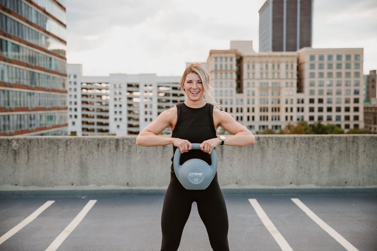 Woman in black workout outfit lifting a kettlebell on a rooftop with city buildings in the background. Photographed by Katie Chamberlain