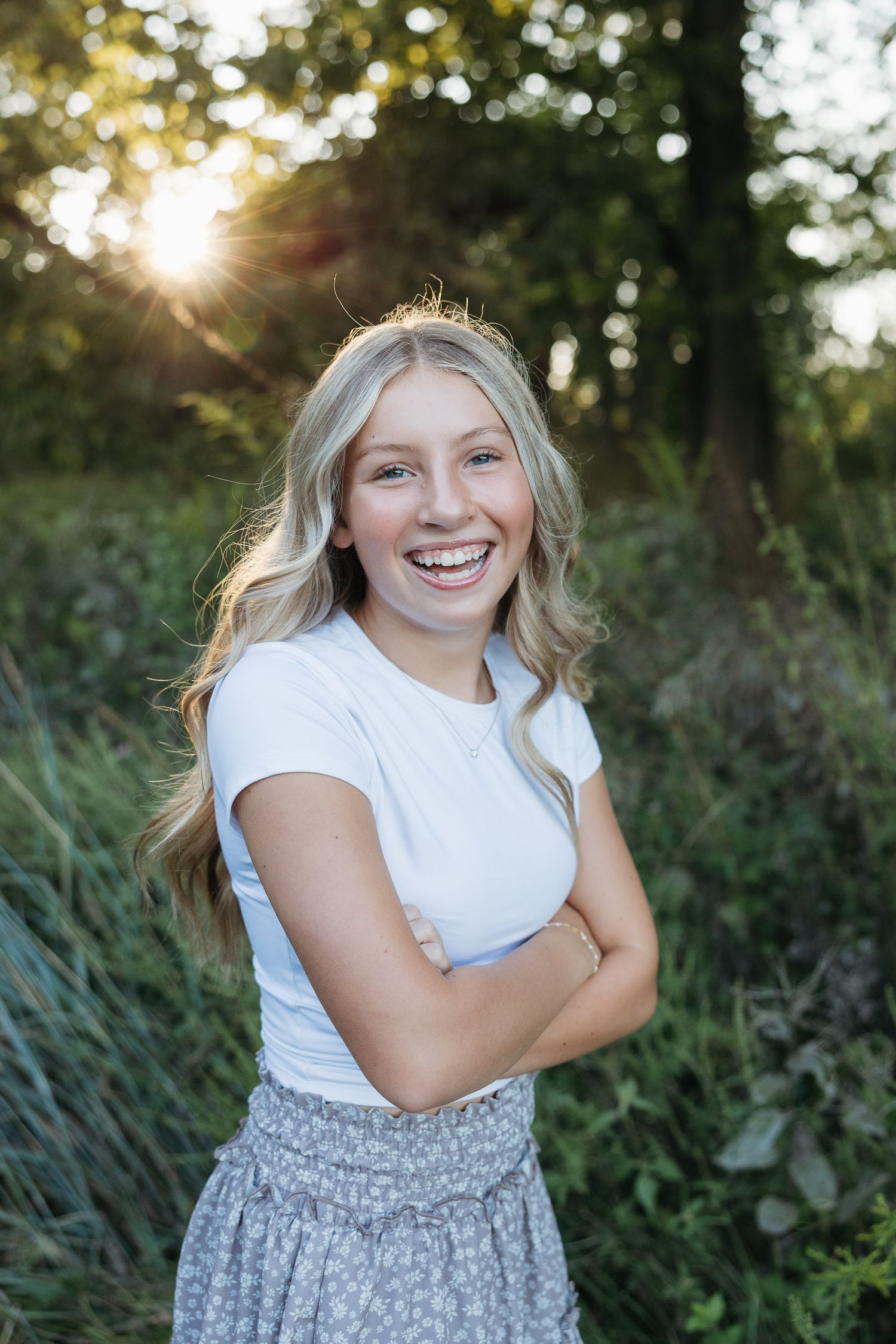 Smiling blonde teenage girl with arms crossed wearing white tee and floral skirt outdoors at golden hour.