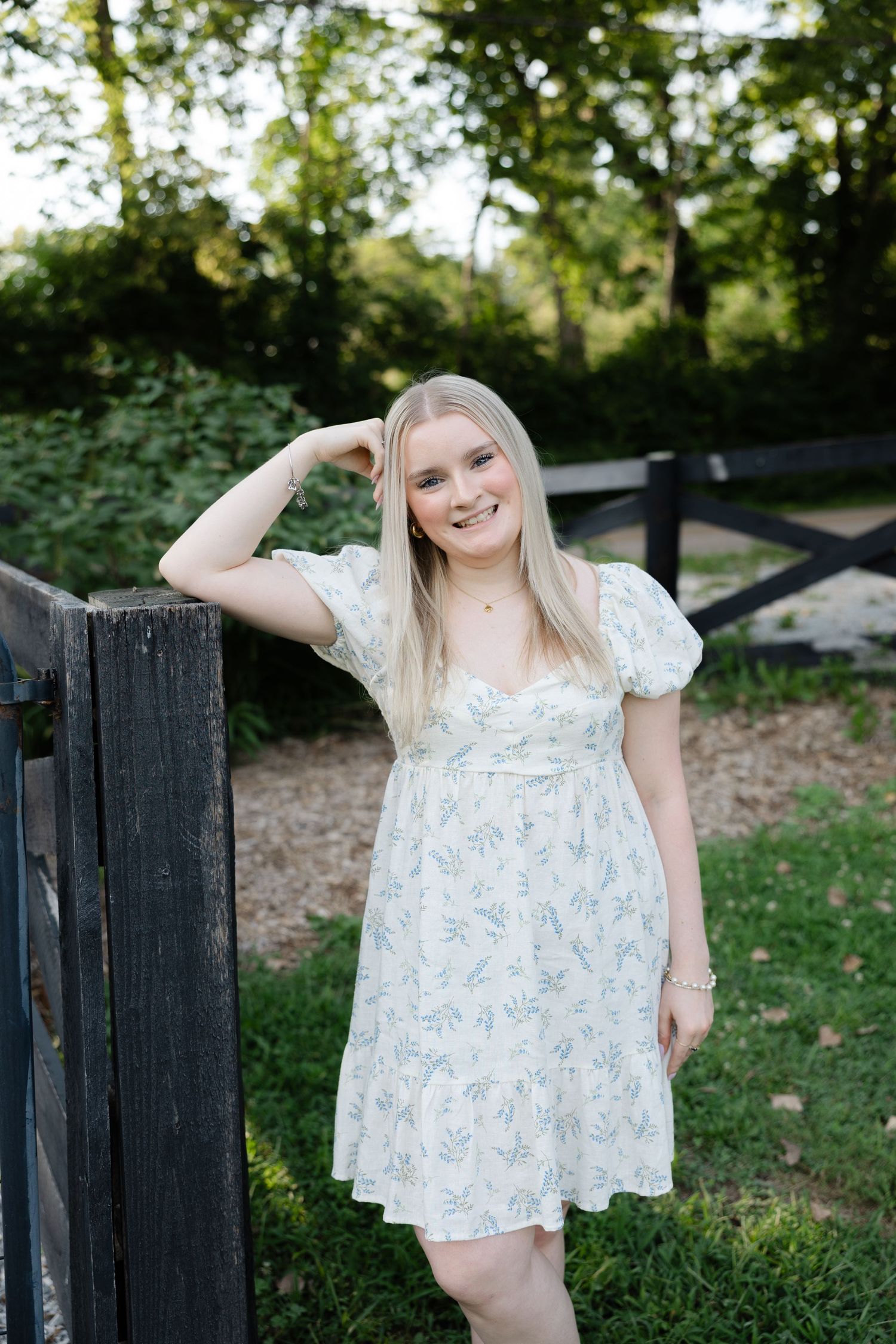 Young blonde woman in floral white dress smiling outdoors near wooden fence with green trees background.
