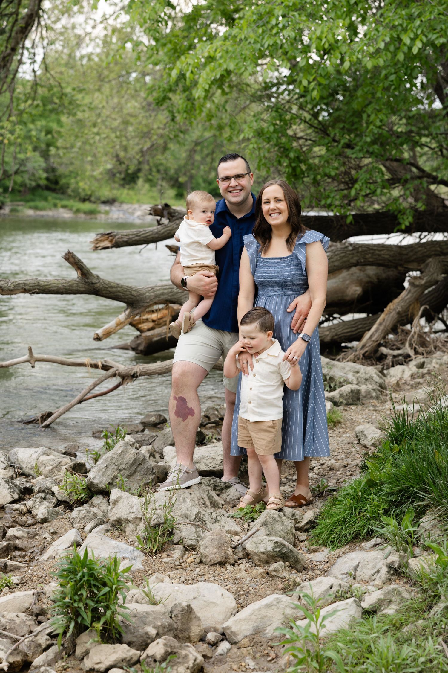 Family of four posing by a rocky riverbank with lush green trees, parents holding young children in a candid outdoor portrait.