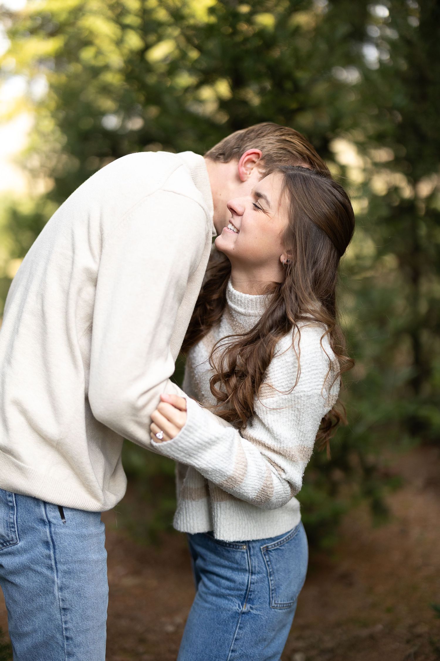 Couple in matching cream sweaters and jeans sharing a tender moment outdoors among green trees. Photographed by Katie Chamberlain Photo