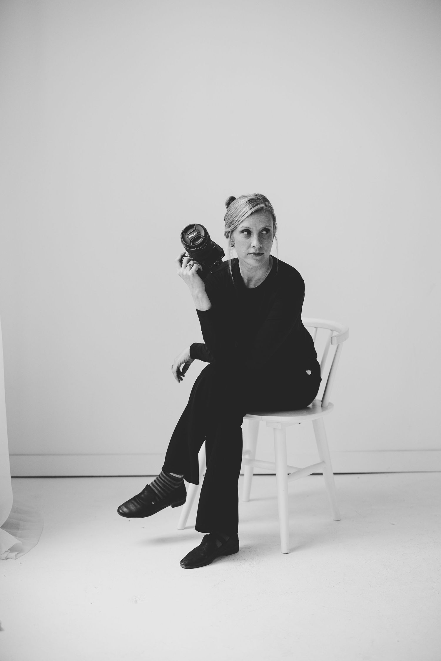 Black and white portrait of a woman in all-black outfit sitting on white chair holding a camera. Katie Chamberlain Photography