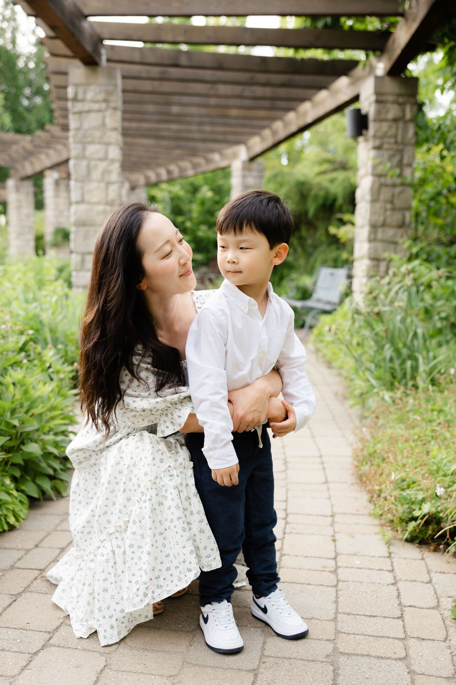 A mother in a floral dress crouches beside her young son in a lush garden pergola walkway.