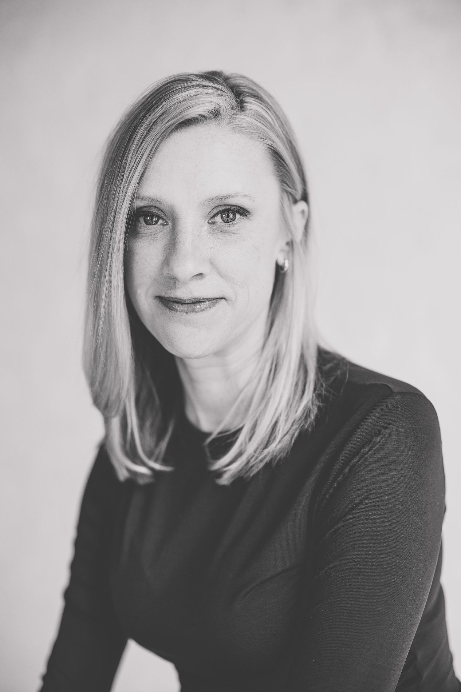 Black and white professional headshot of a blonde woman in a dark top, smiling confidently at the camera. Katie Chamberlain Photography
