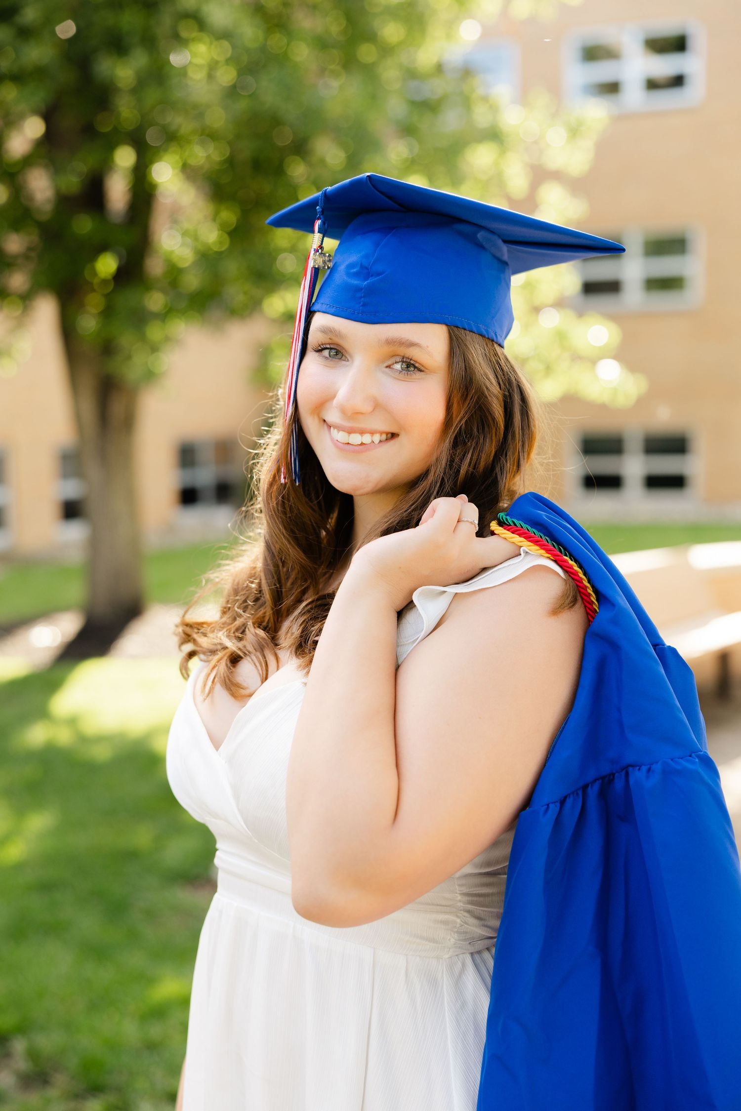 Smiling female graduate in blue cap and gown holding diploma outside school building on sunny day.