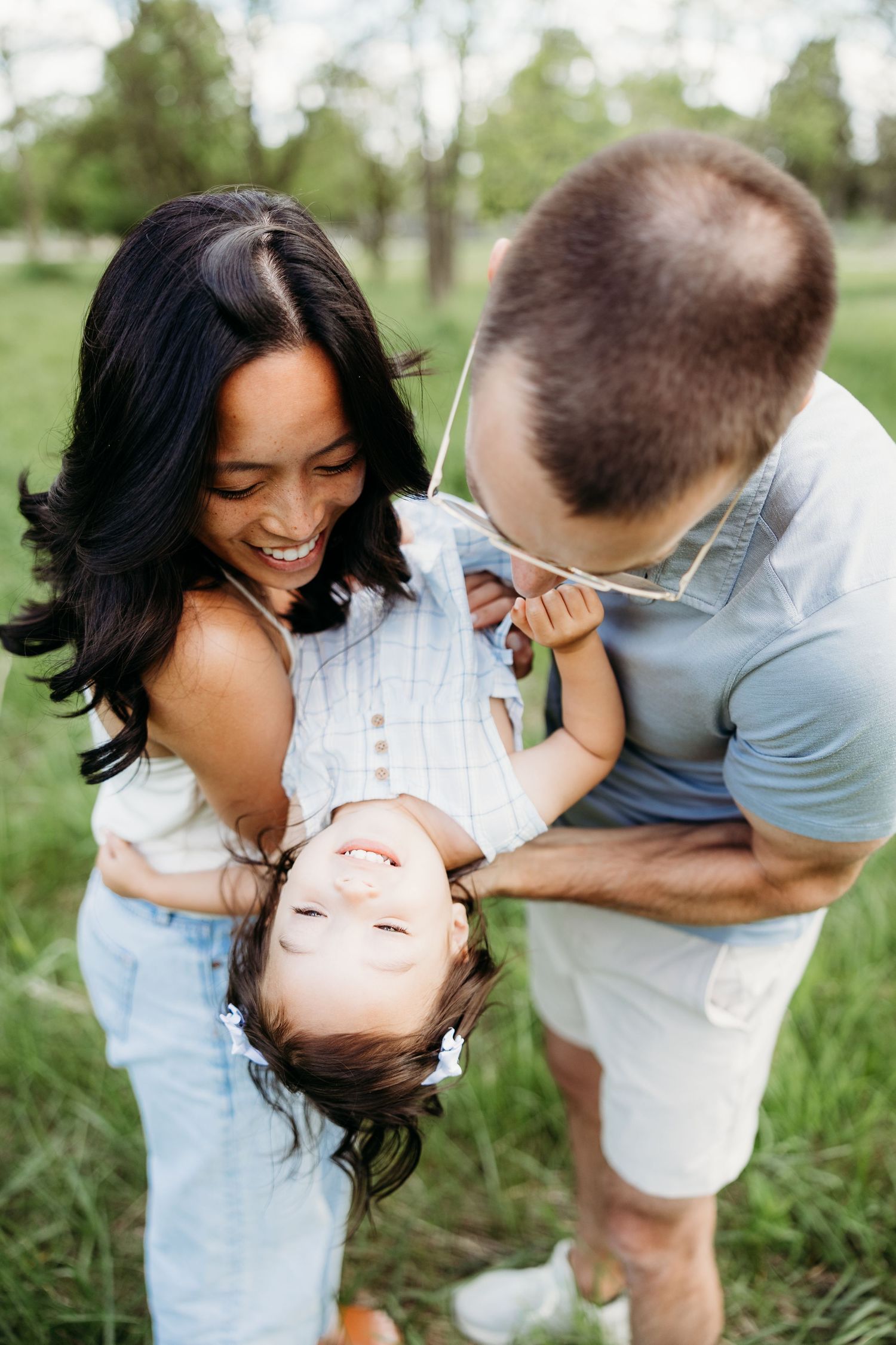 Happy family playing outdoors, parents laughing while holding young child upside down in green park setting.