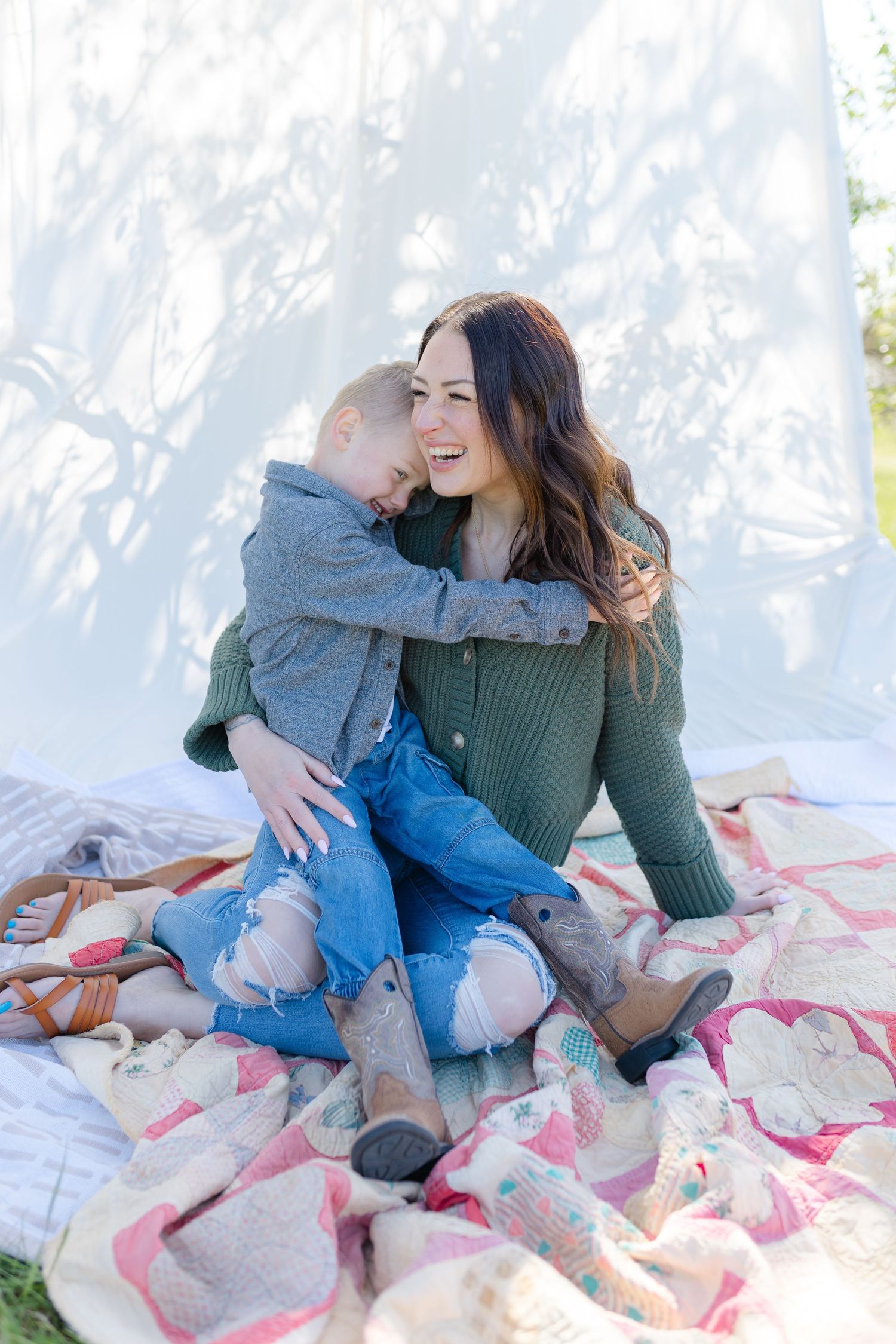 A laughing woman in a green sweater and jeans sits on a quilt outdoors while a young child hugs her affectionately.