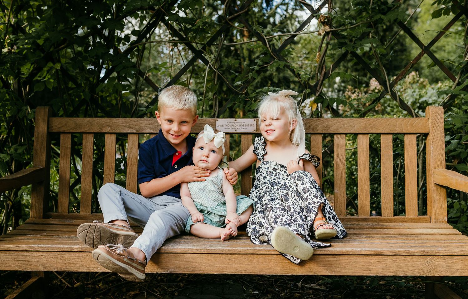 Three young children sitting together on a wooden garden bench surrounded by green foliage. Photographed by Katie Chamberlain Photography