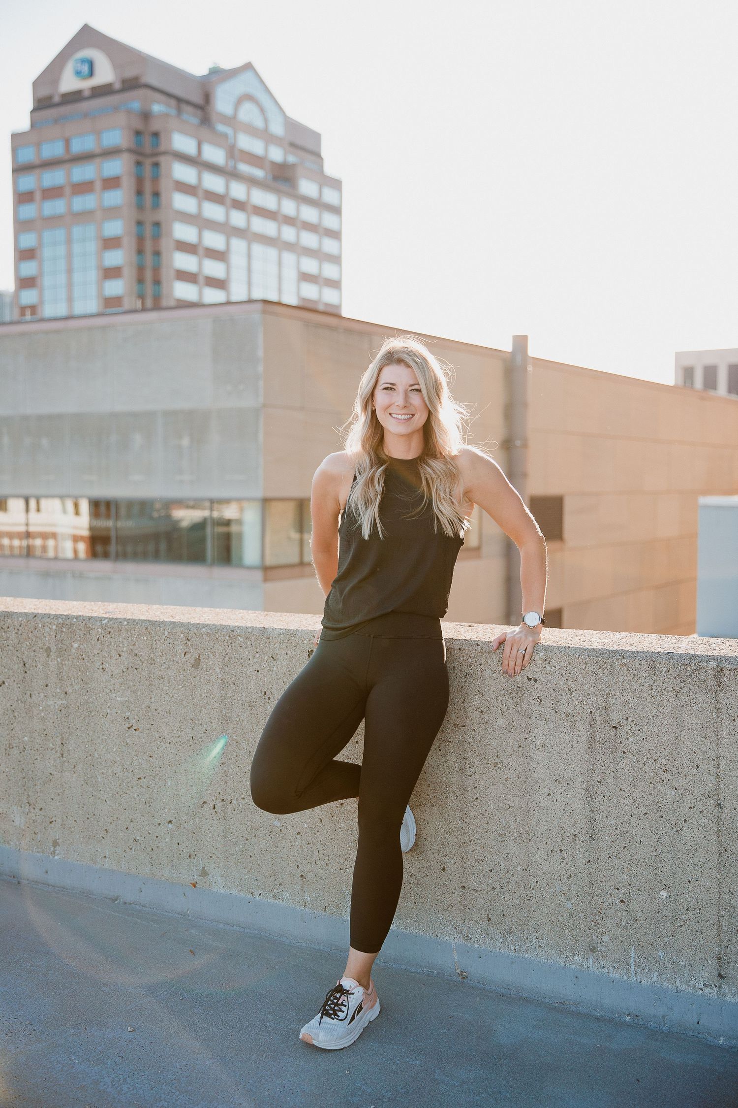 Smiling blonde woman in black athletic wear leaning against a concrete wall urban buildings in the background. Photo by Katie Chamberlain